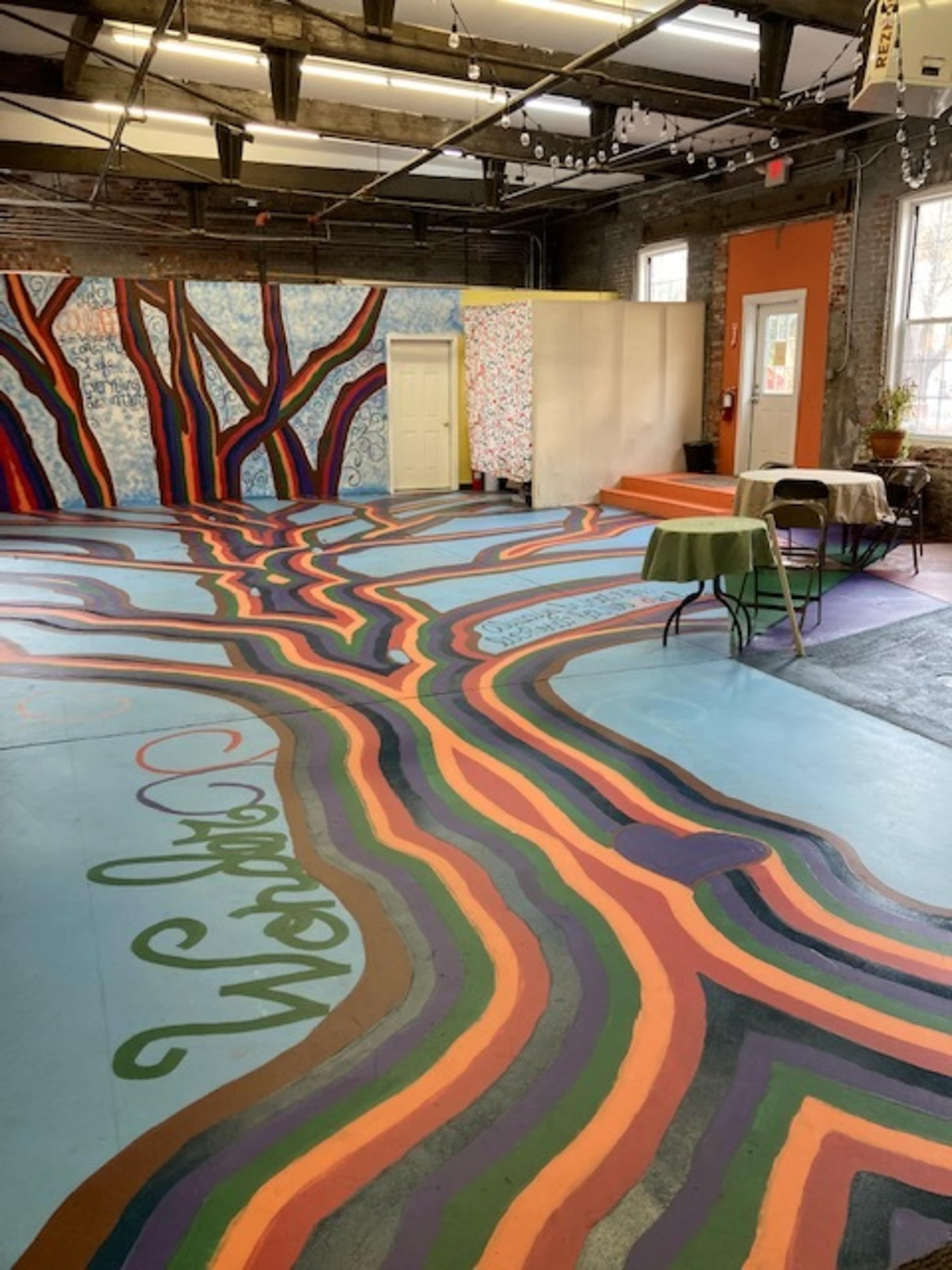 A vibrant, mural-adorned room with painted tree patterns on the floor and colorful walls, featuring a few tables arranged in the space.