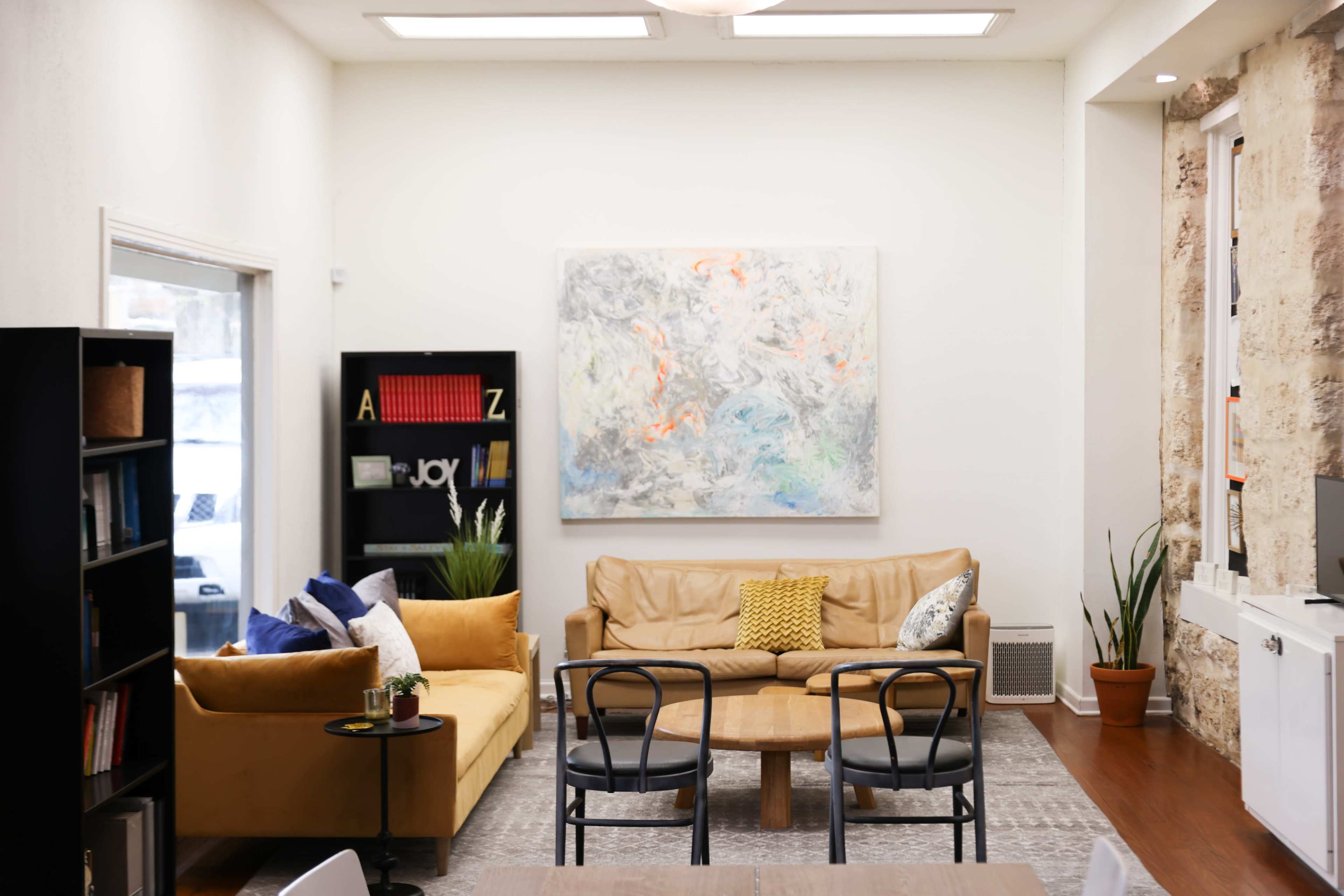 A cozy lounge area features two sofas, a coffee table, and a large abstract painting on the wall, alongside bookshelves and potted plants.