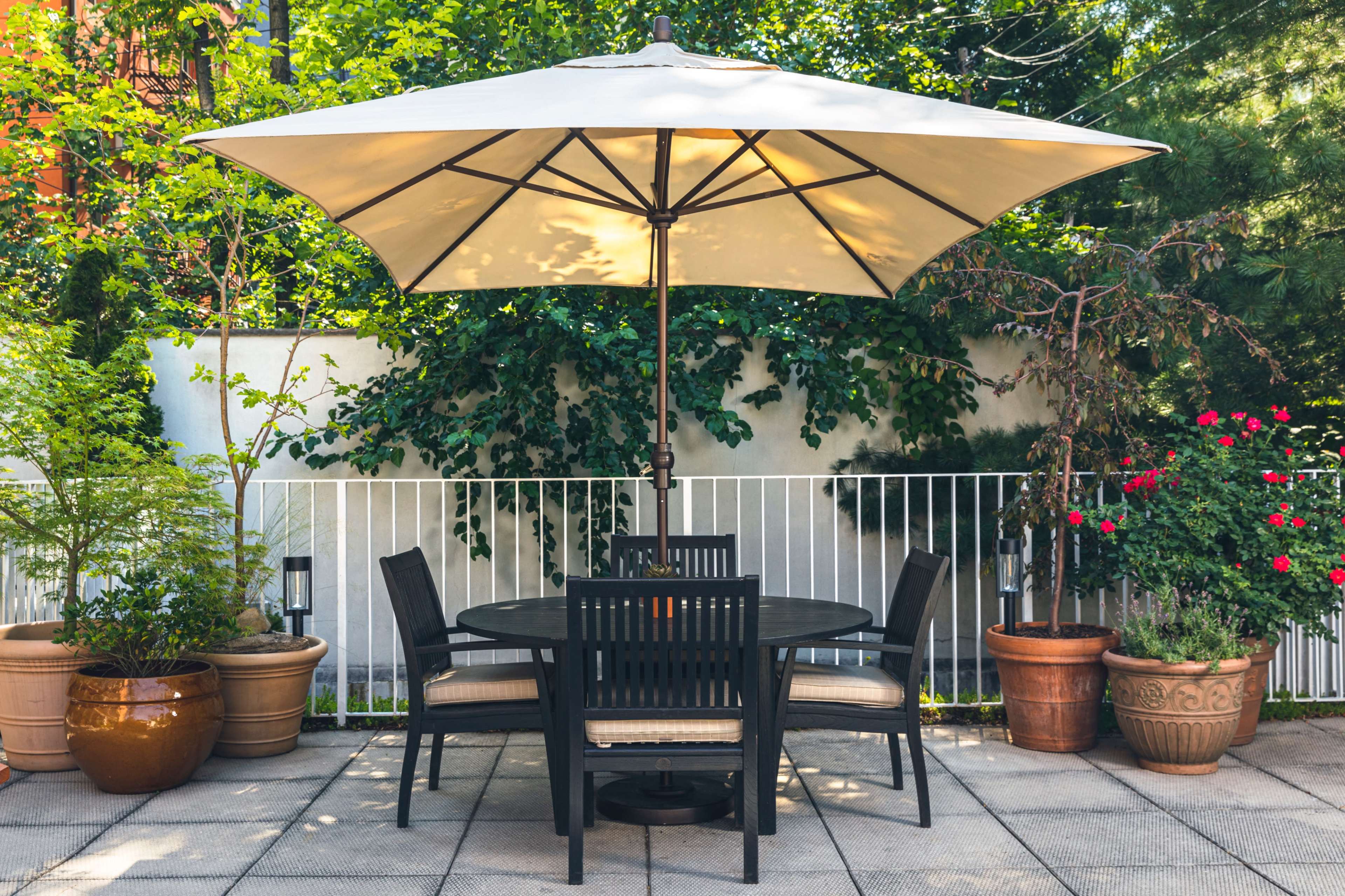 A round black table with four chairs is situated under a large cream-colored umbrella on a paved patio surrounded by greenery and potted plants.