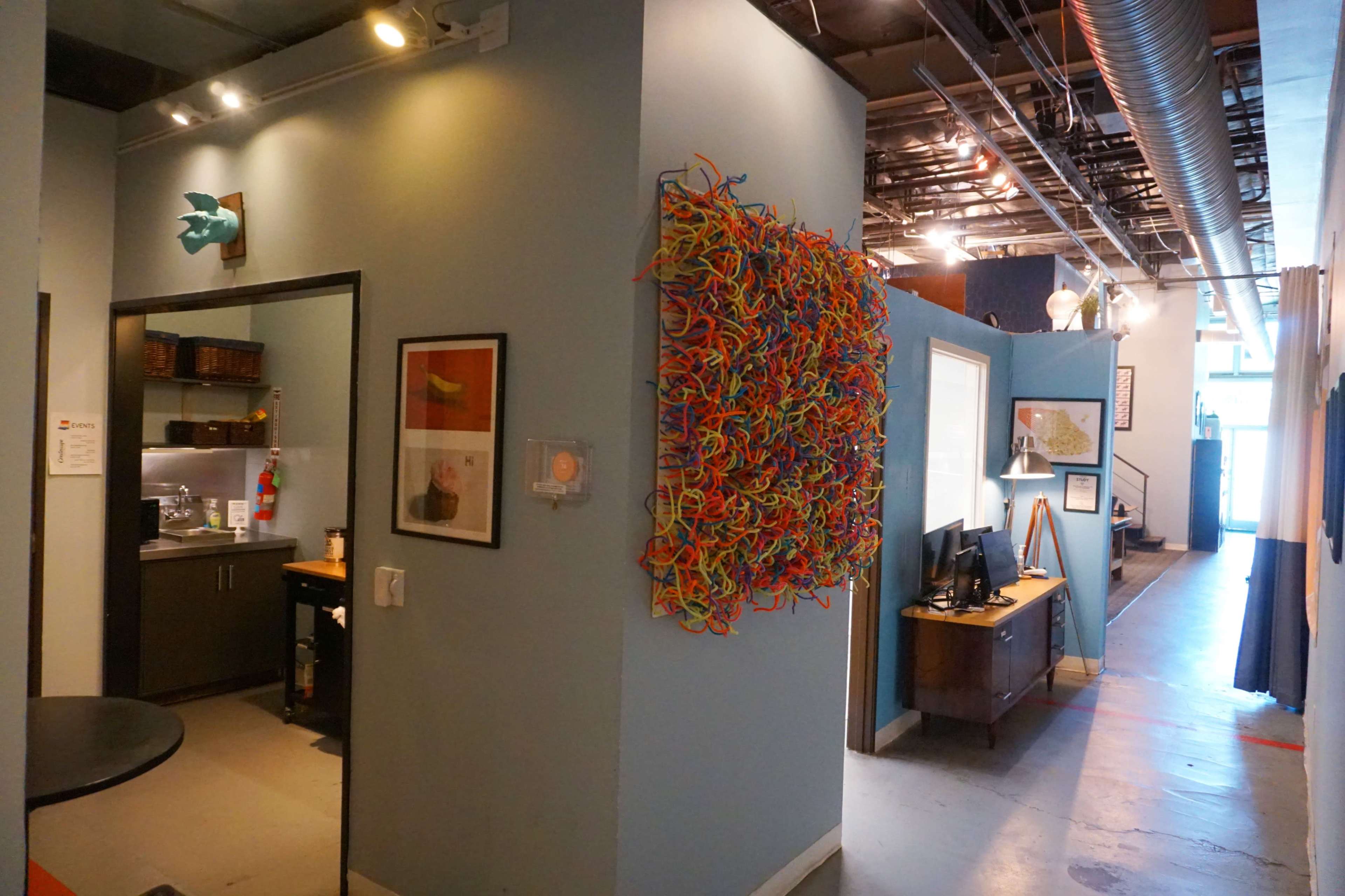 The image shows a hallway with a colorful artwork made of intertwined plastic strands on one wall, leading to a kitchen area and an office setup.