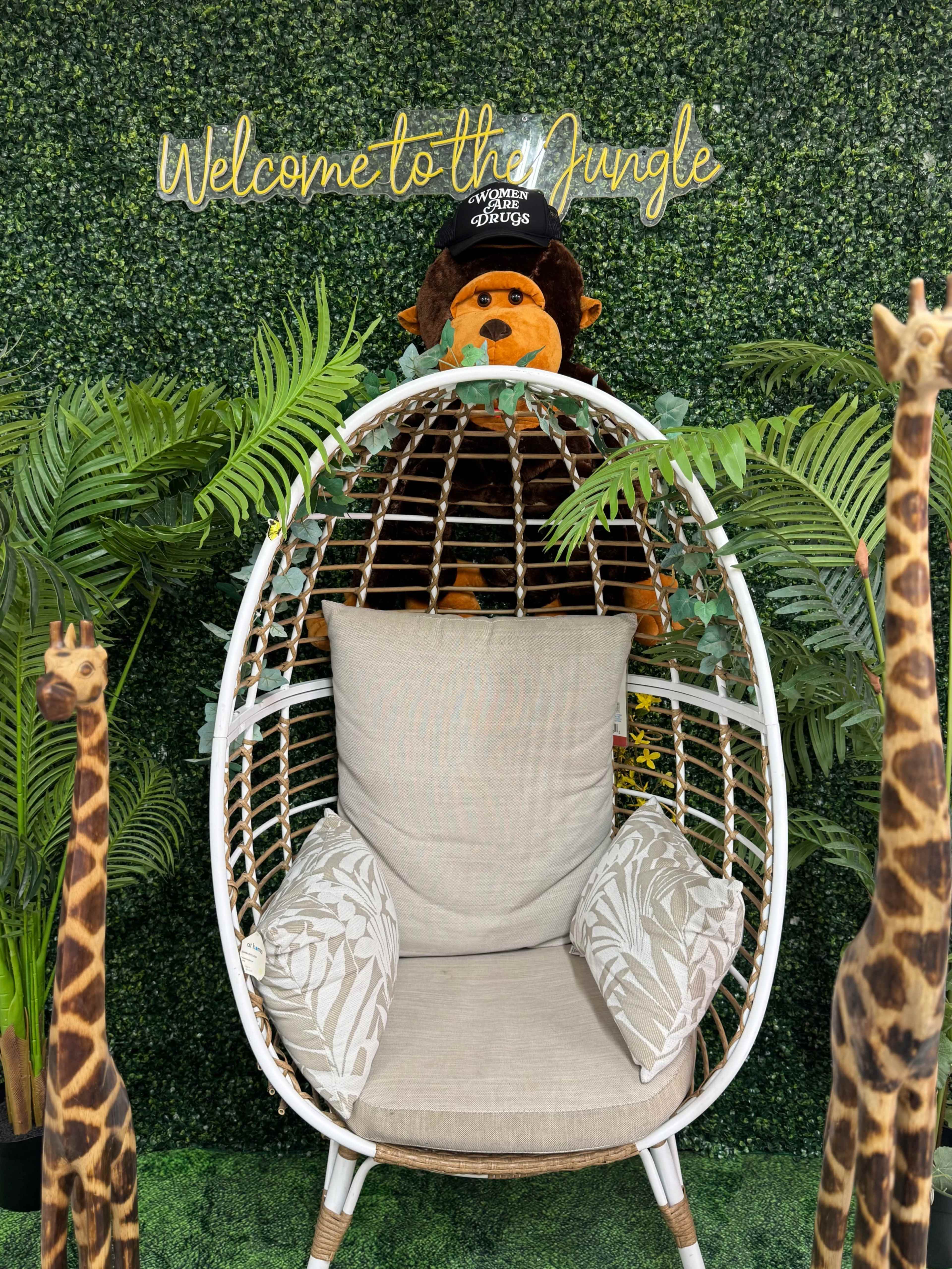 A white hanging chair surrounded by faux greenery and two giraffe statues, with a stuffed monkey placed on top.