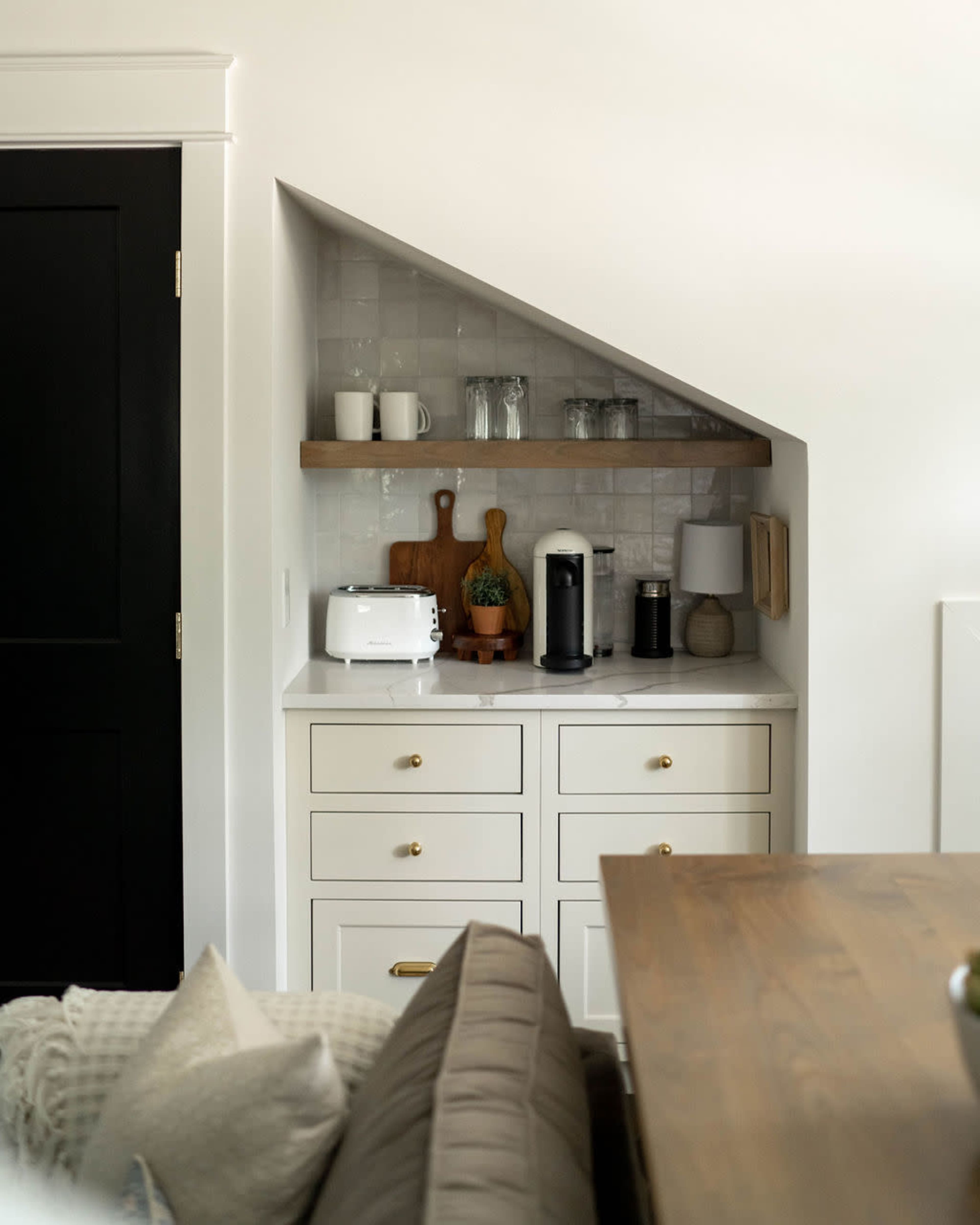 A compact kitchen nook features a toaster, coffee maker, and glassware arranged on a countertop below a sloped wall with a shelf.