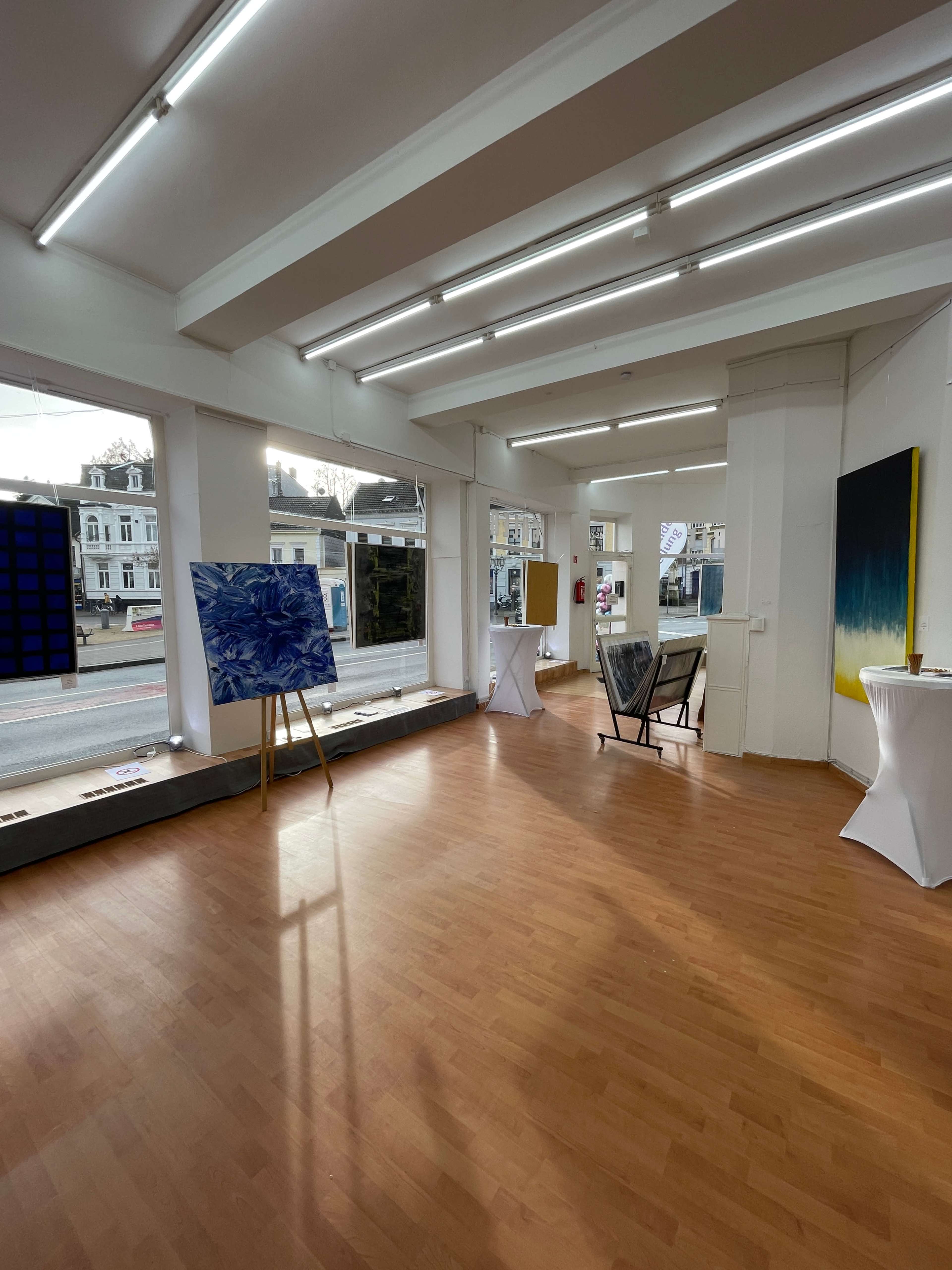 A spacious art gallery with wooden flooring and walls displaying various artworks in a well-lit setting.