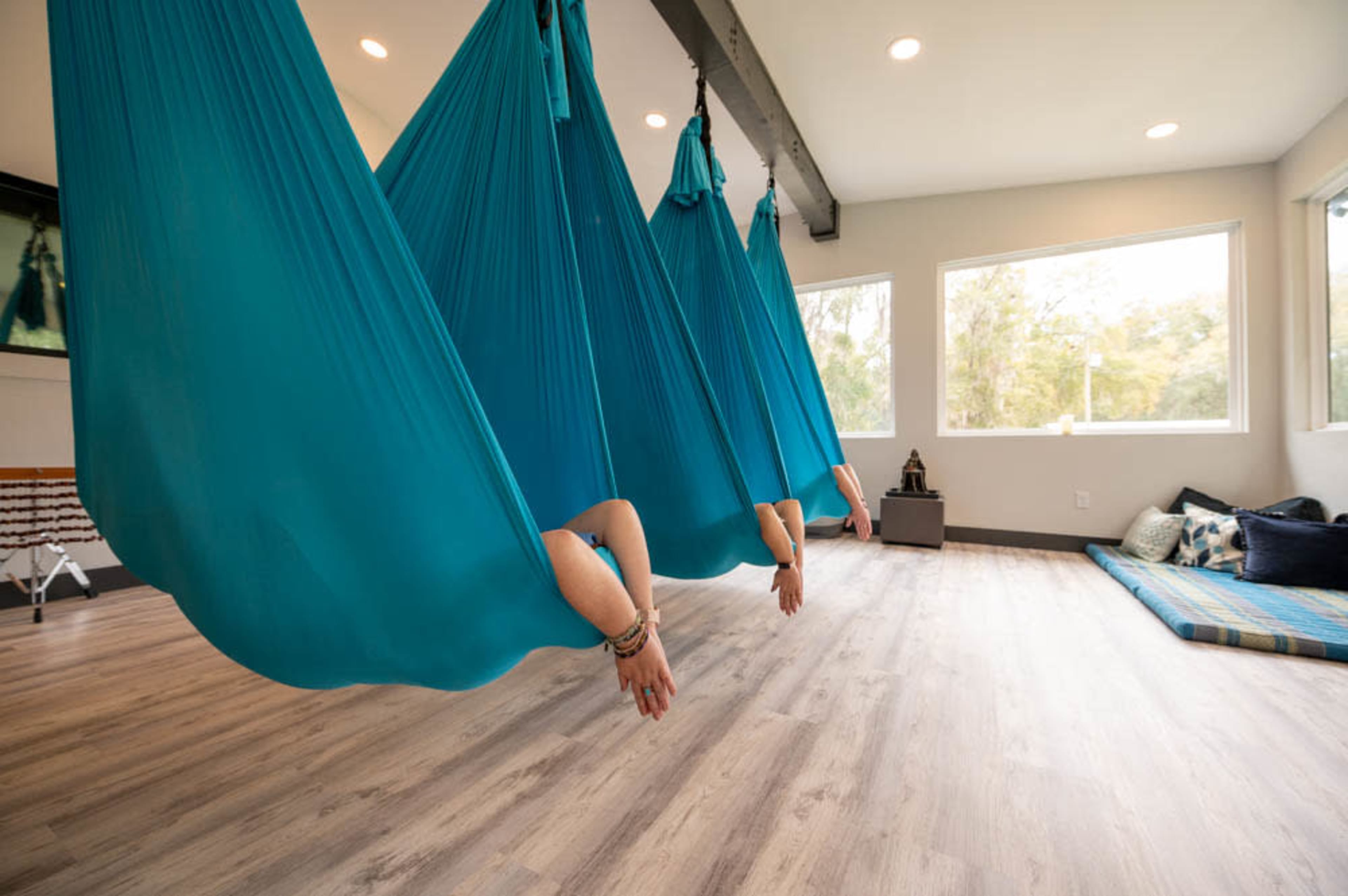 Three suspended aerial silks hang from the ceiling in a bright room with large windows and a wooden floor.