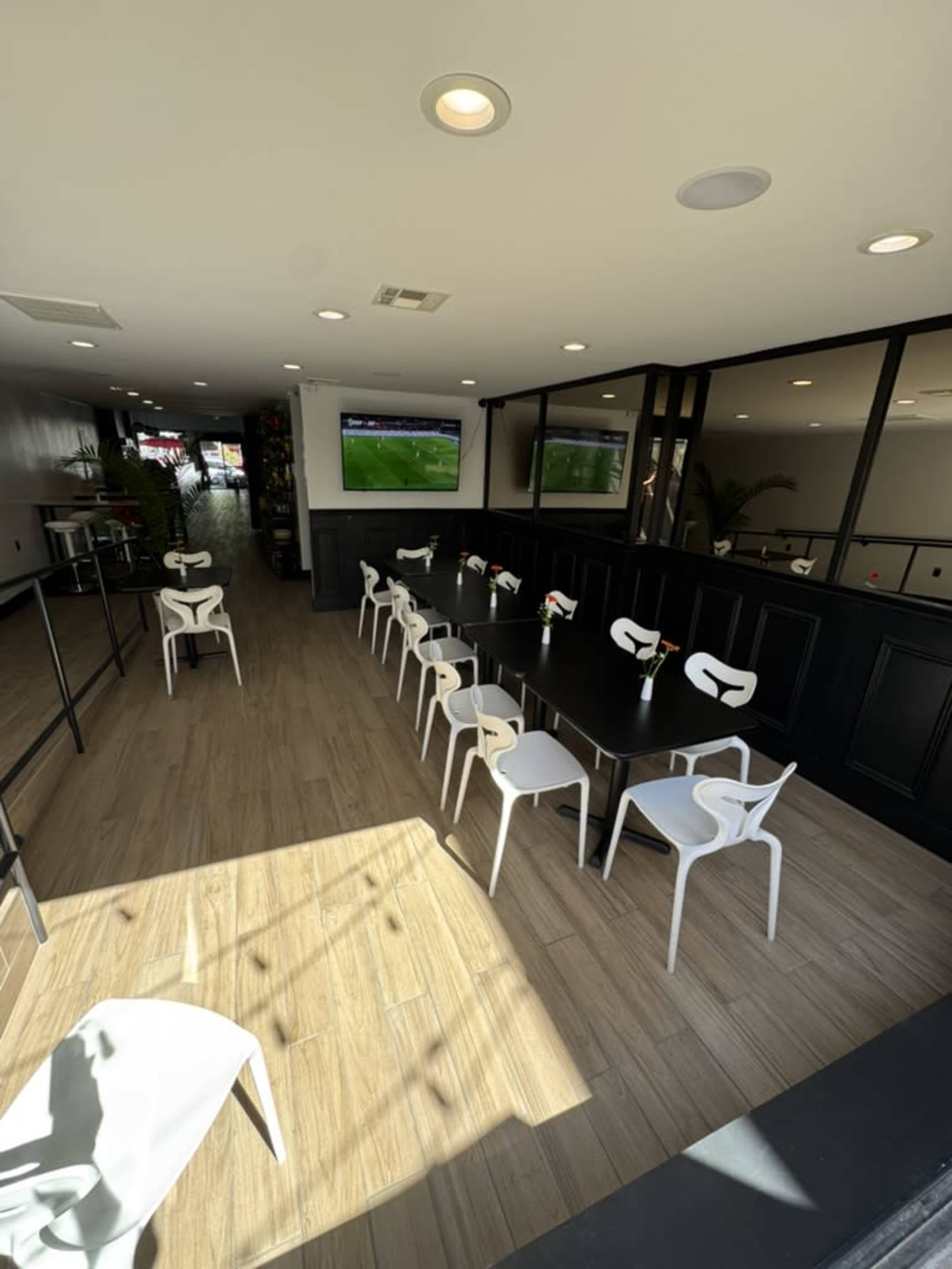The image shows a modern, well-lit dining area with a long black table surrounded by white chairs, and a large screen displaying a sports event.