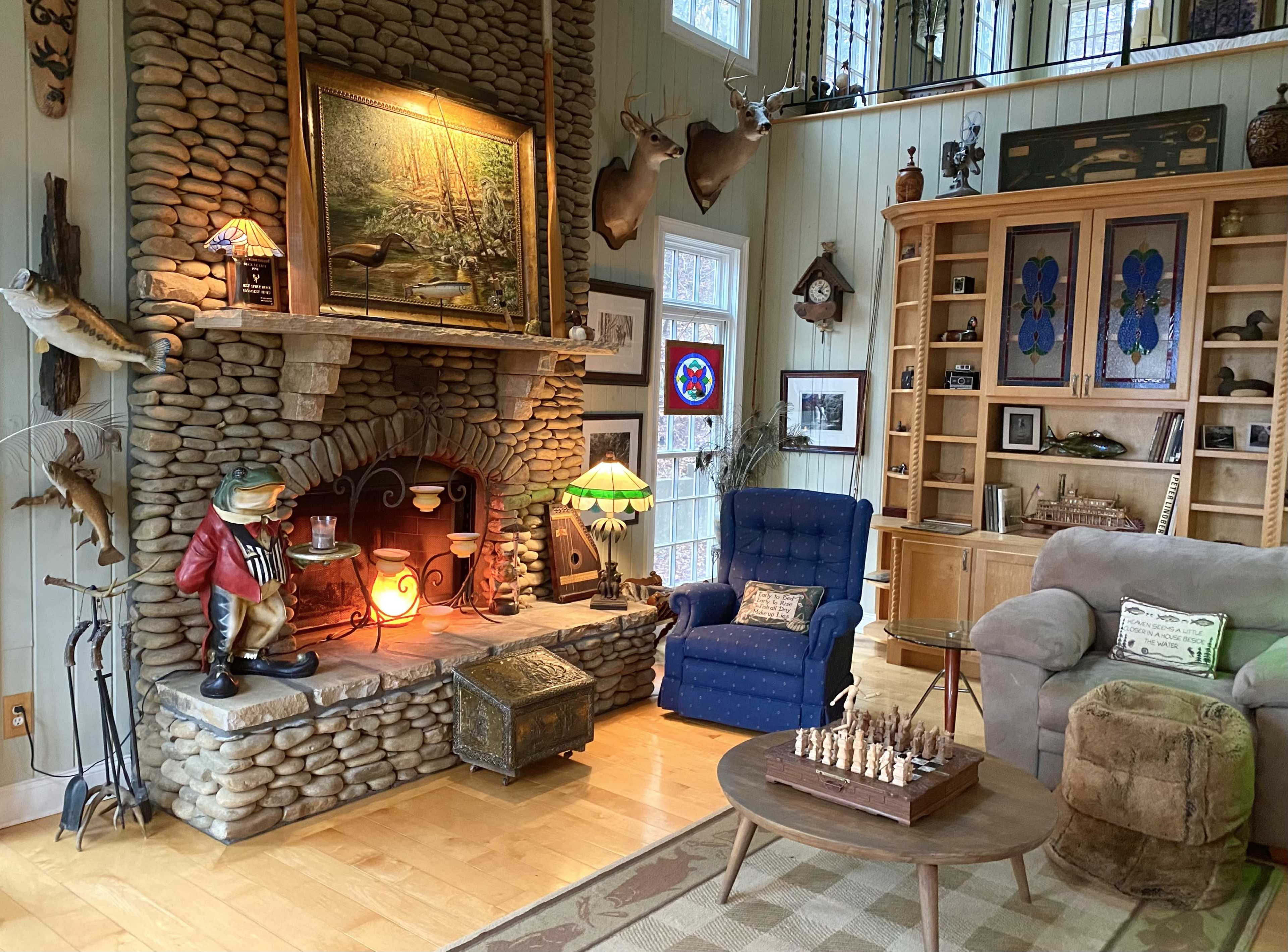 The room features a stone fireplace with a rustic decor, including mounted deer heads, a wooden chess set on a round table, and various collectibles displayed on shelves.