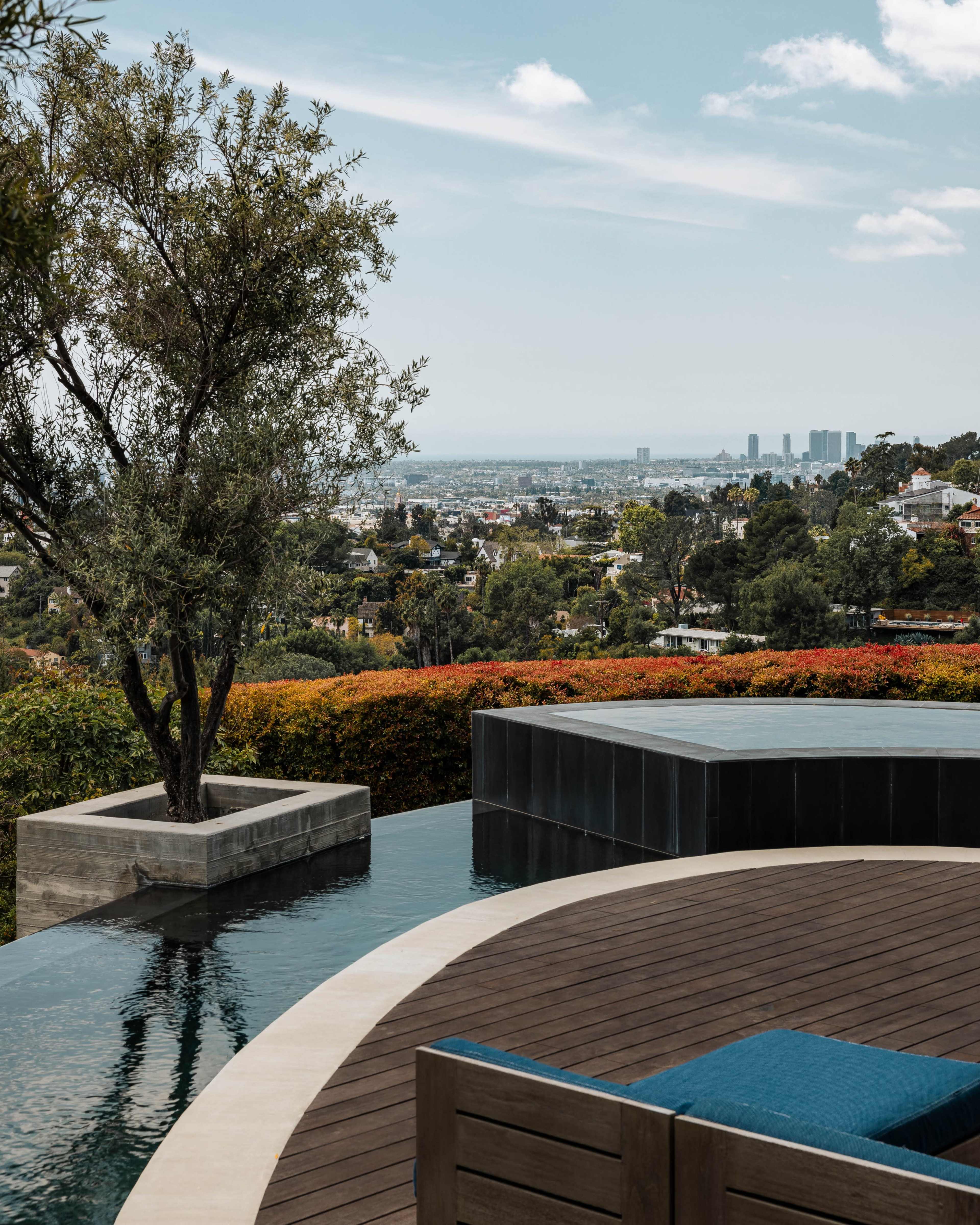 The image shows a modern outdoor space with a circular wooden deck, a small tree beside a pool, and a panoramic view of a city in the distance.