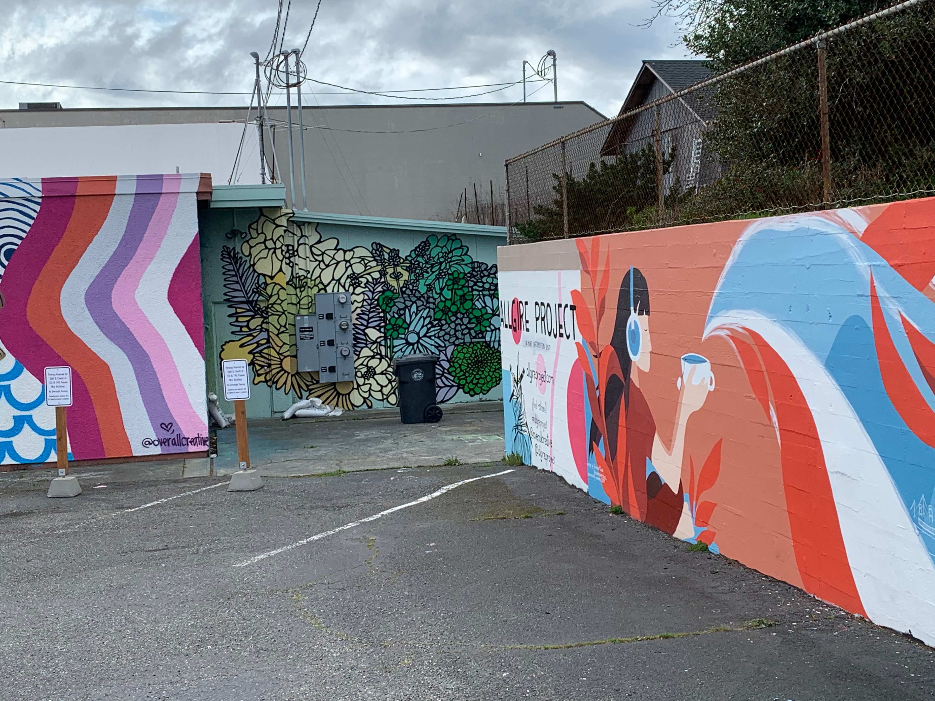 A parking area featuring colorful murals on adjacent walls, with a trash bin and utility equipment in the background.