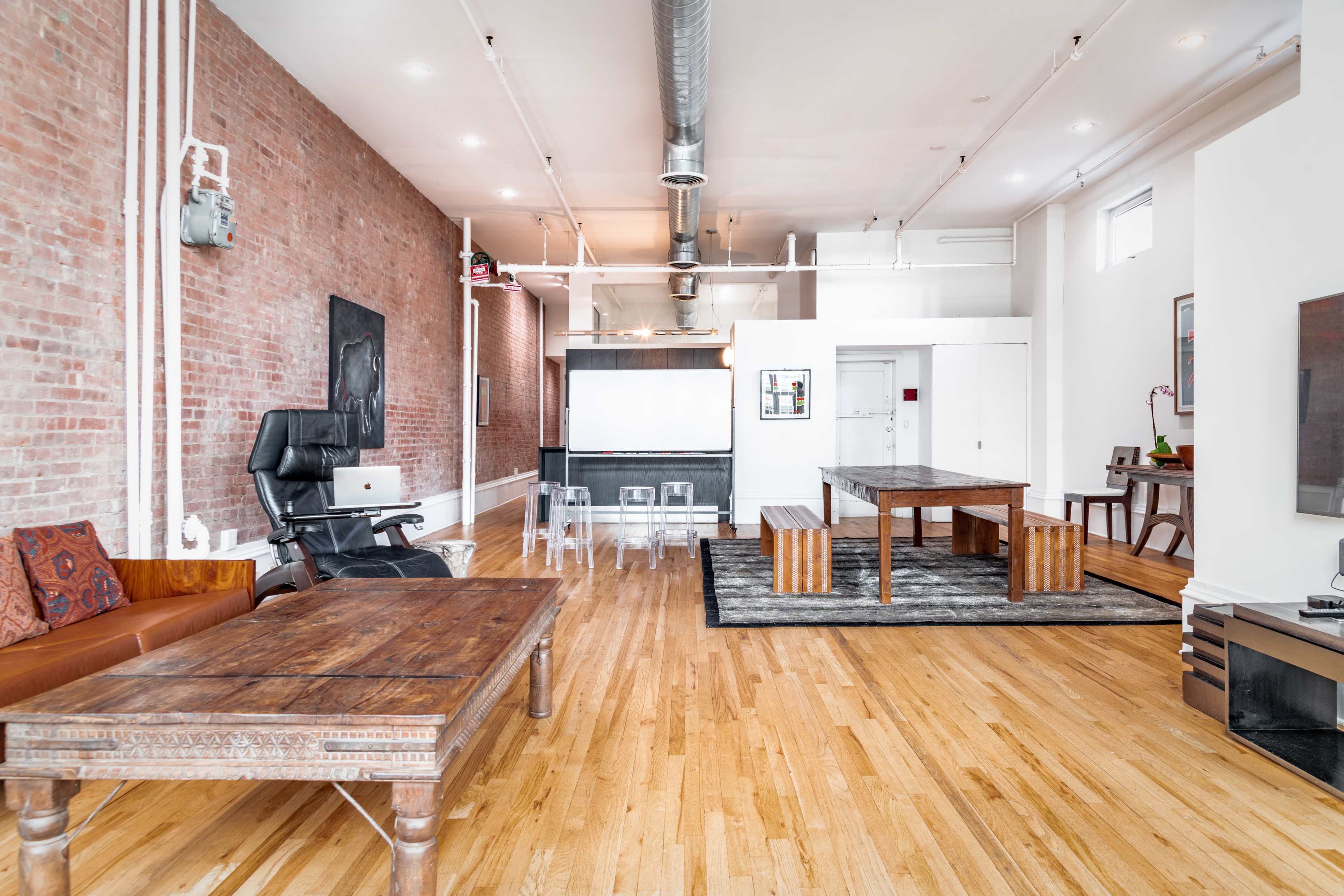 The image shows a spacious, modern living area with wooden flooring, a large dining table, a dark kitchen area in the background, and a combination of rustic and contemporary furniture.