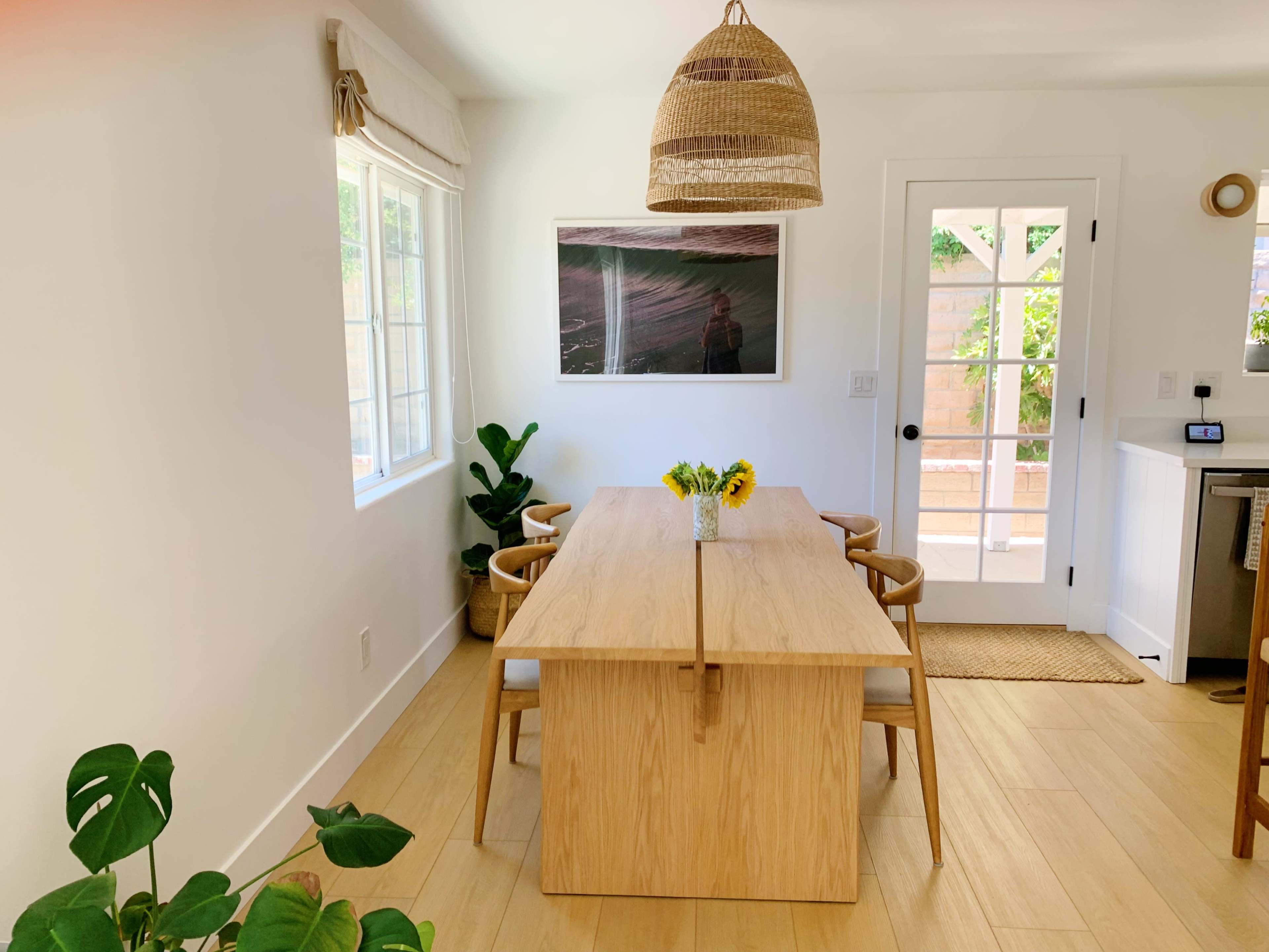 A wooden dining table with chairs is centered in a sunlit room that features a large artwork on the wall and plants in the corners.