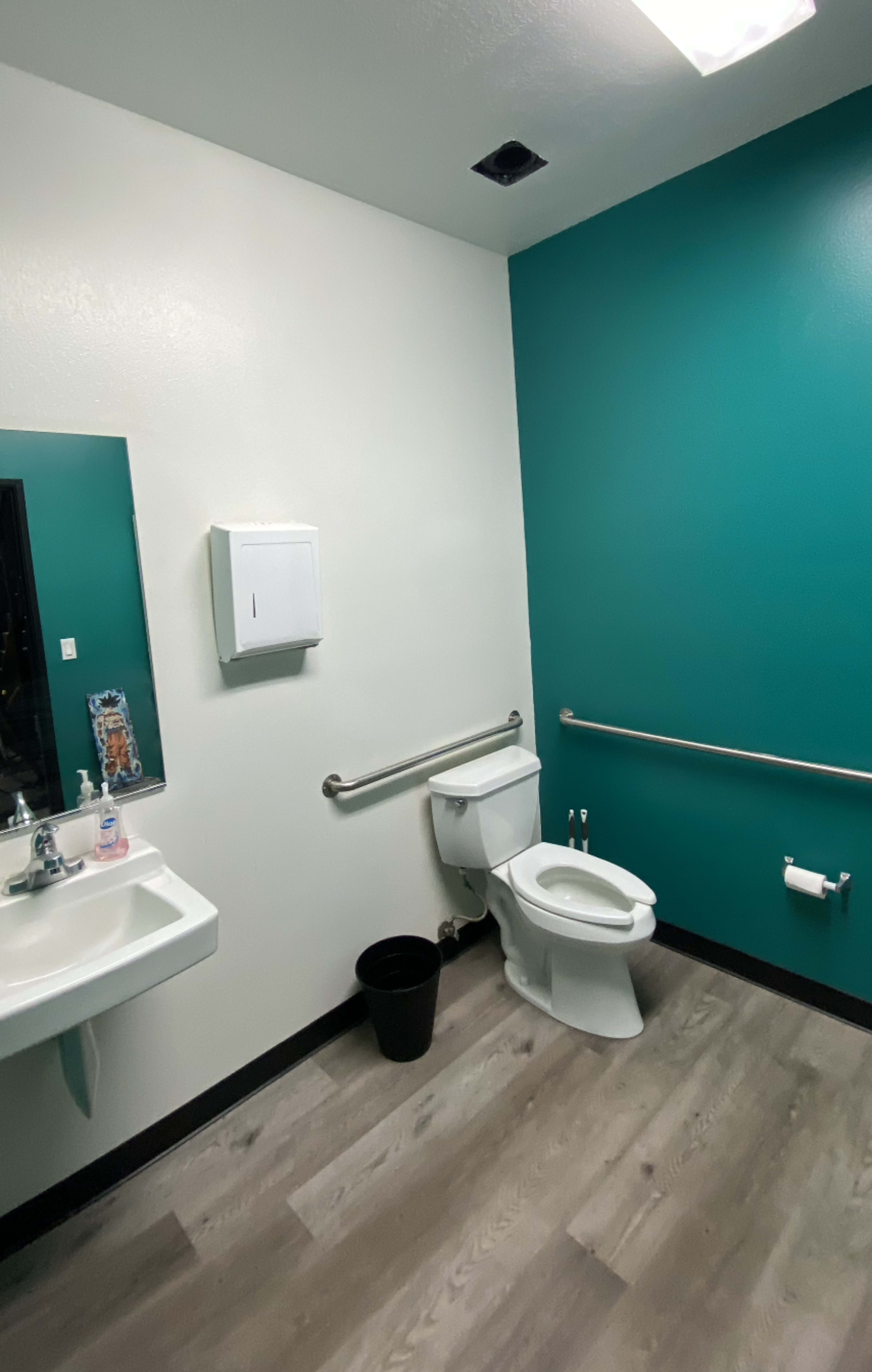 A restroom with a toilet, a sink, and teal walls, featuring a large mirror and one paper towel dispenser.