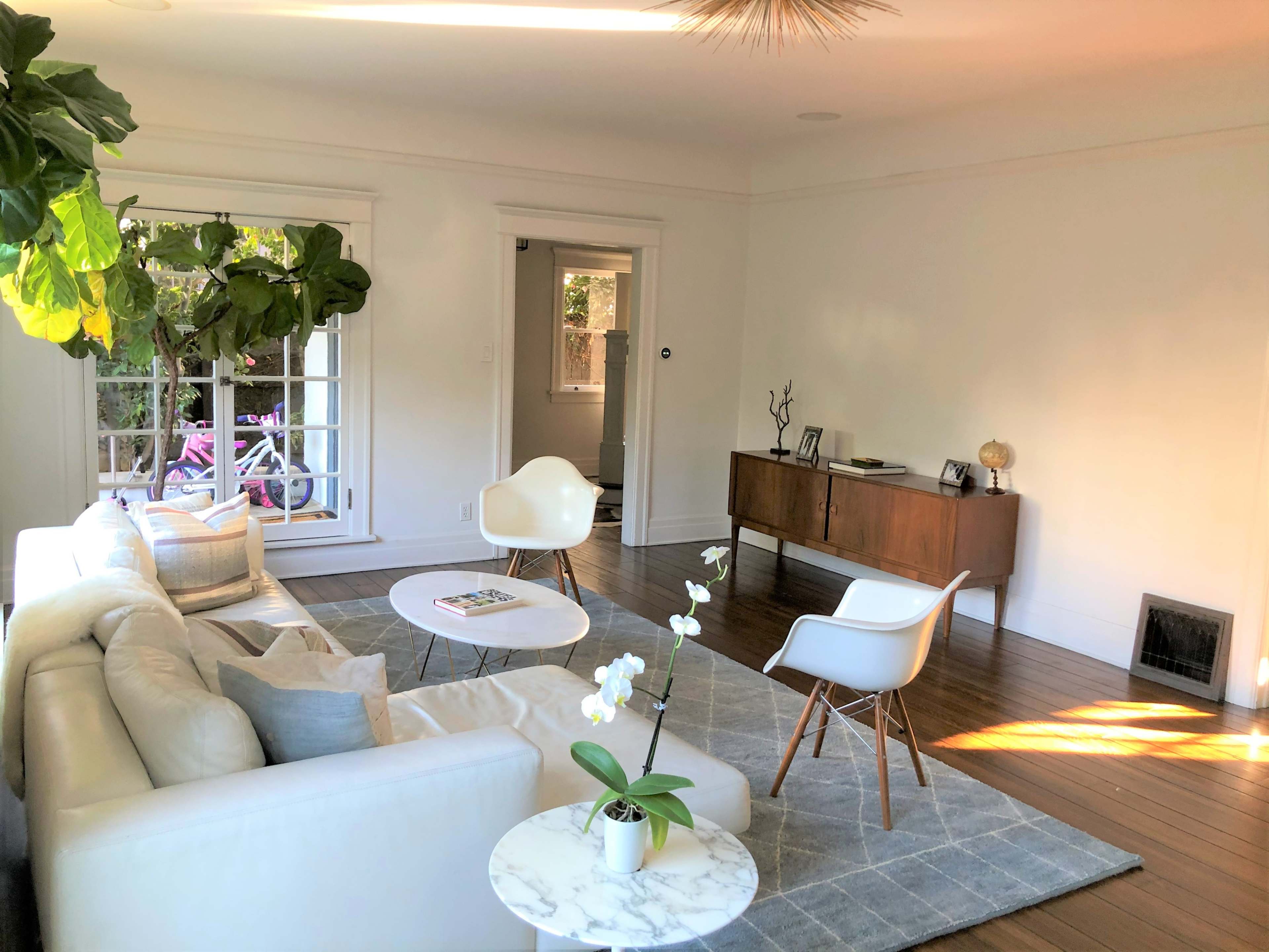 The scene depicts a bright living room with a white couch, a round coffee table, and a wooden sideboard, featuring plants and neutral decor on the walls.