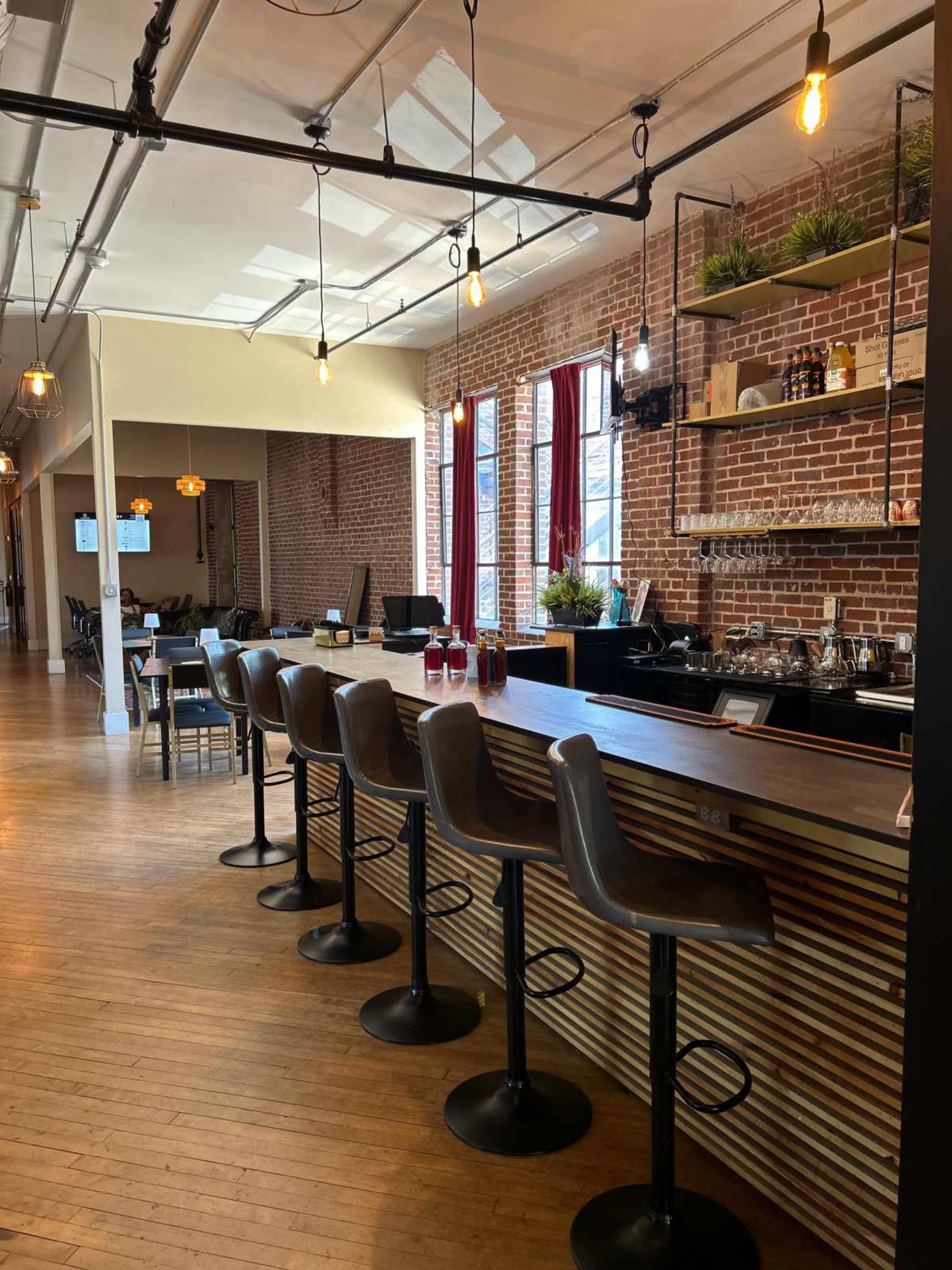 The image shows a modern bar area featuring high stools along a wooden counter, with exposed brick walls and large windows providing natural light.