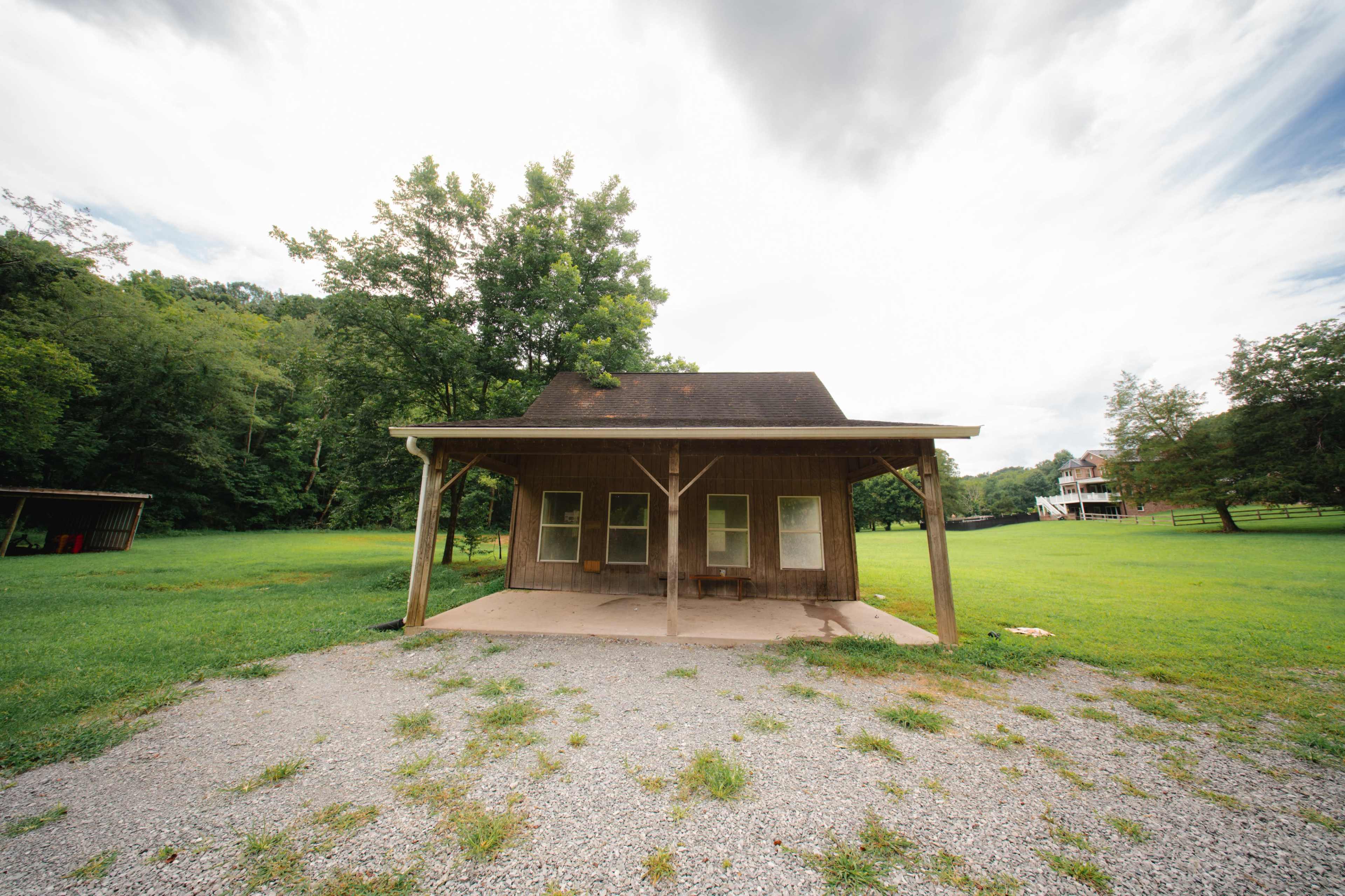A small, wooden structure with a porch is situated on a grassy area surrounded by trees, with clouds visible in the sky.