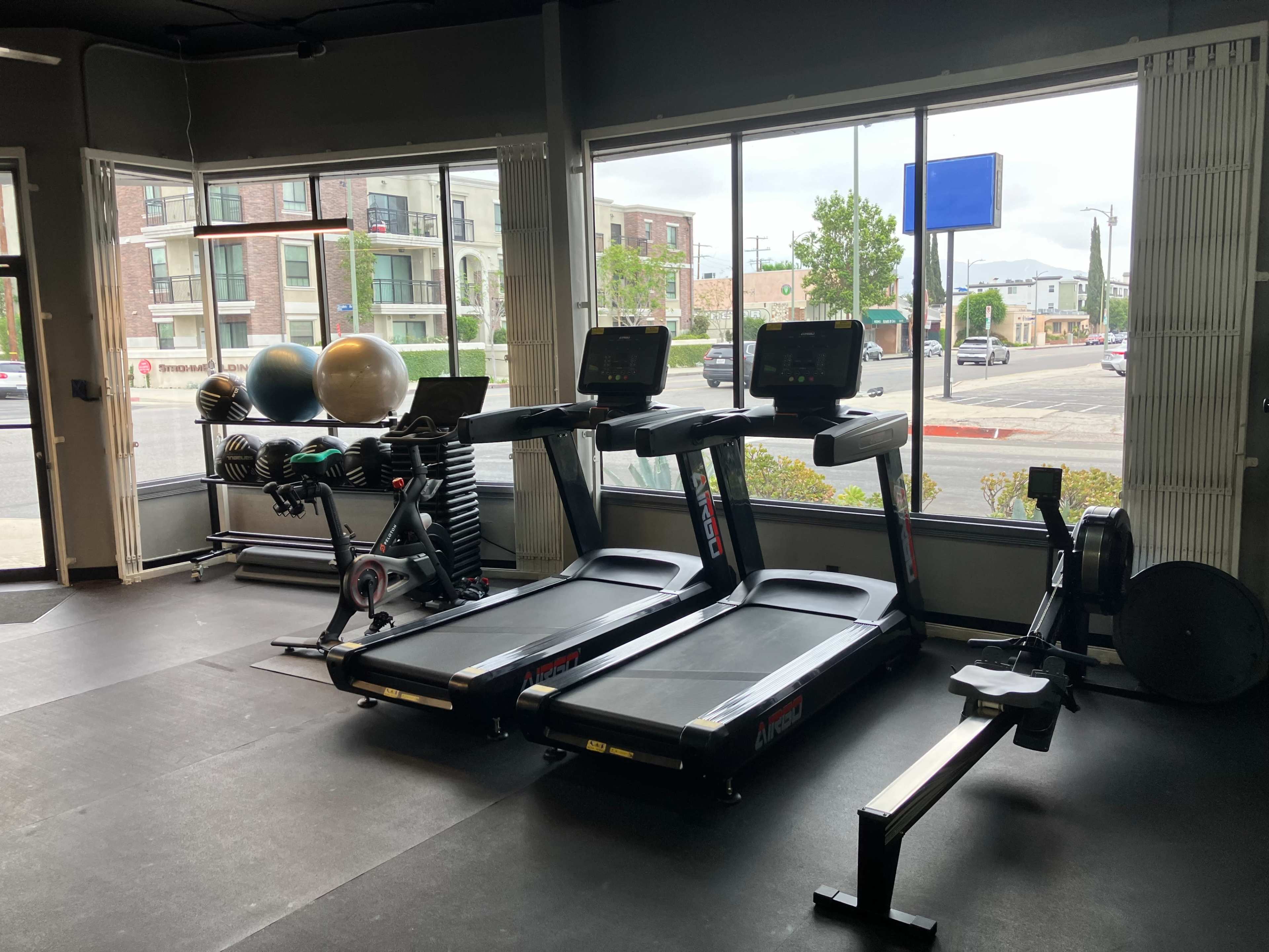 The image shows a fitness area featuring three treadmills, a rowing machine, and exercise equipment arranged near large windows with a view of a street outside.