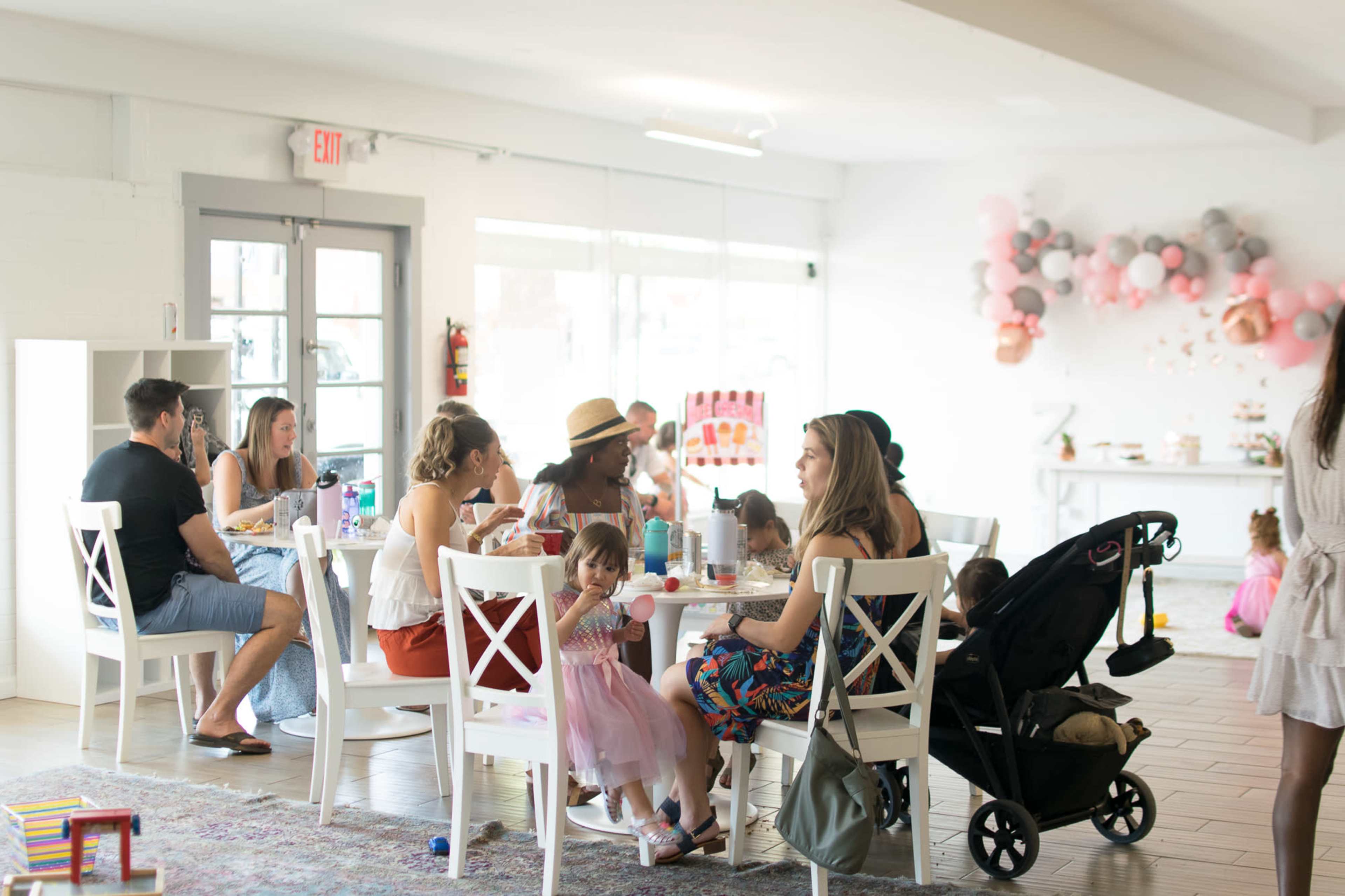 A group of adults and children are seated around tables in a bright, modern event space decorated with balloons.