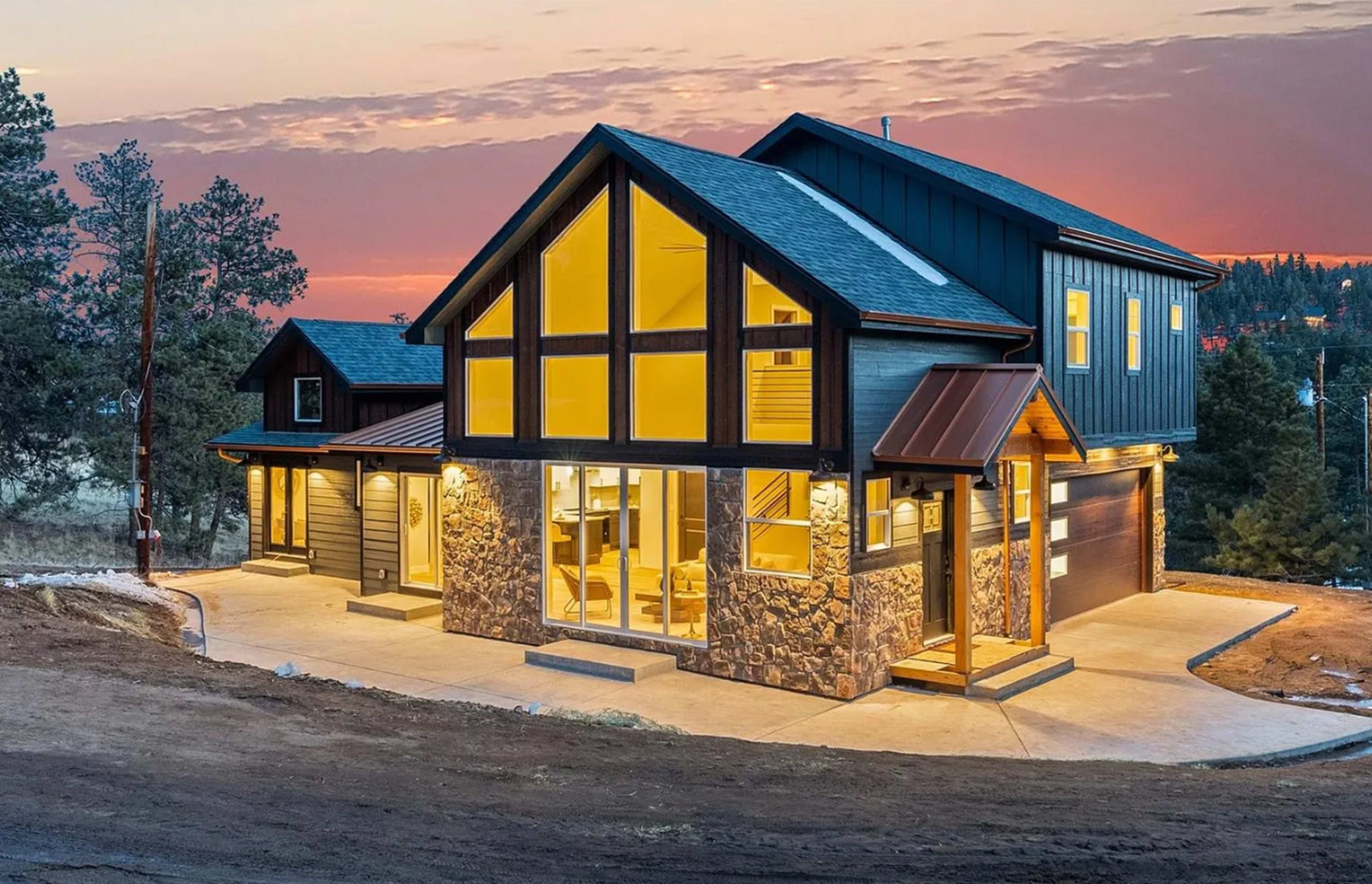 A modern two-story house features a large glass front, stone accents, and a paved driveway, set against a twilight sky.