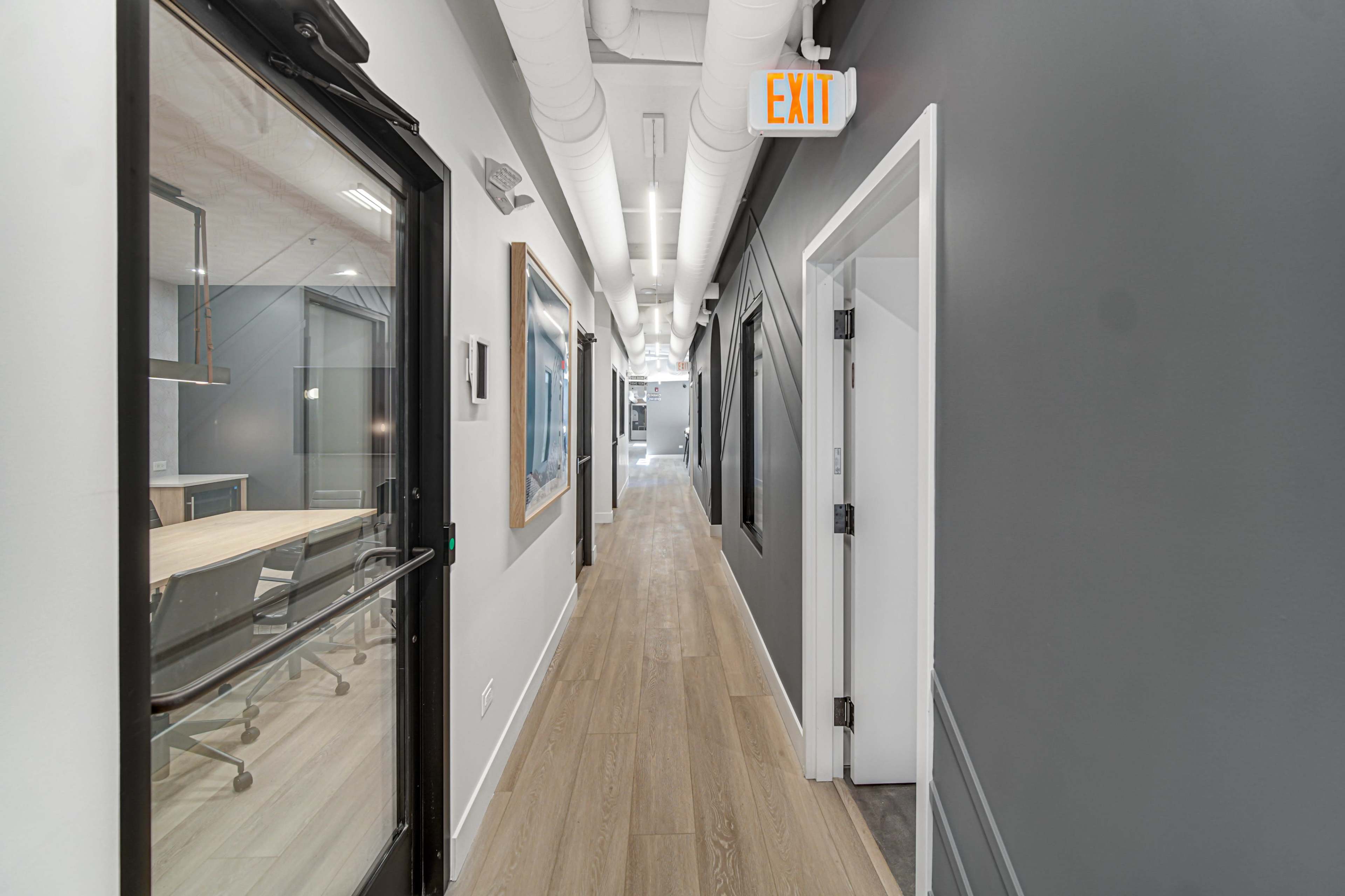 The image shows a narrow hallway in an office building, featuring light-colored walls, wooden flooring, and exit signs.