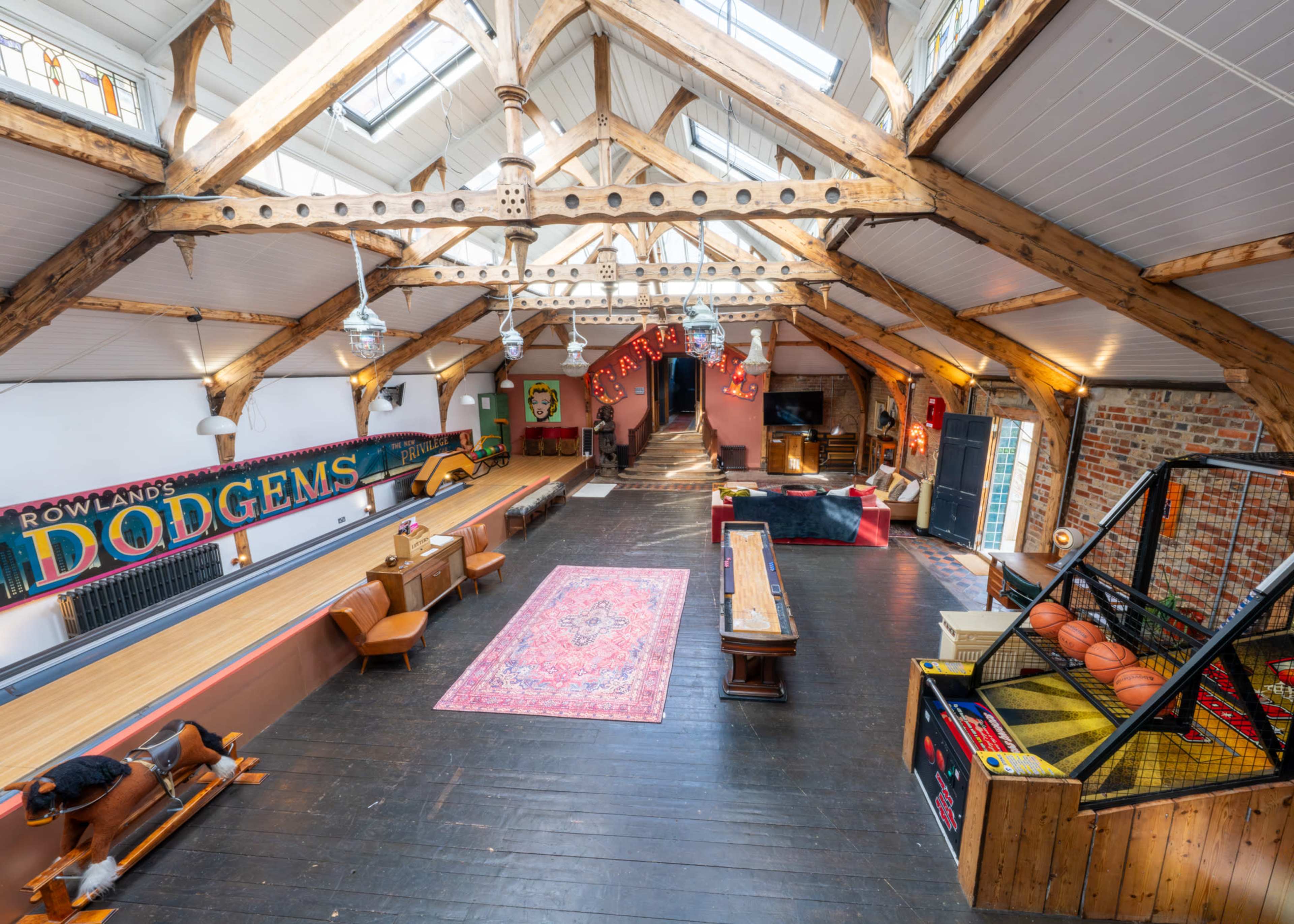 The image shows a spacious interior with exposed wooden beams, featuring a vintage bowling alley, a pool table, and arcade games, all set against a brick wall backdrop.
