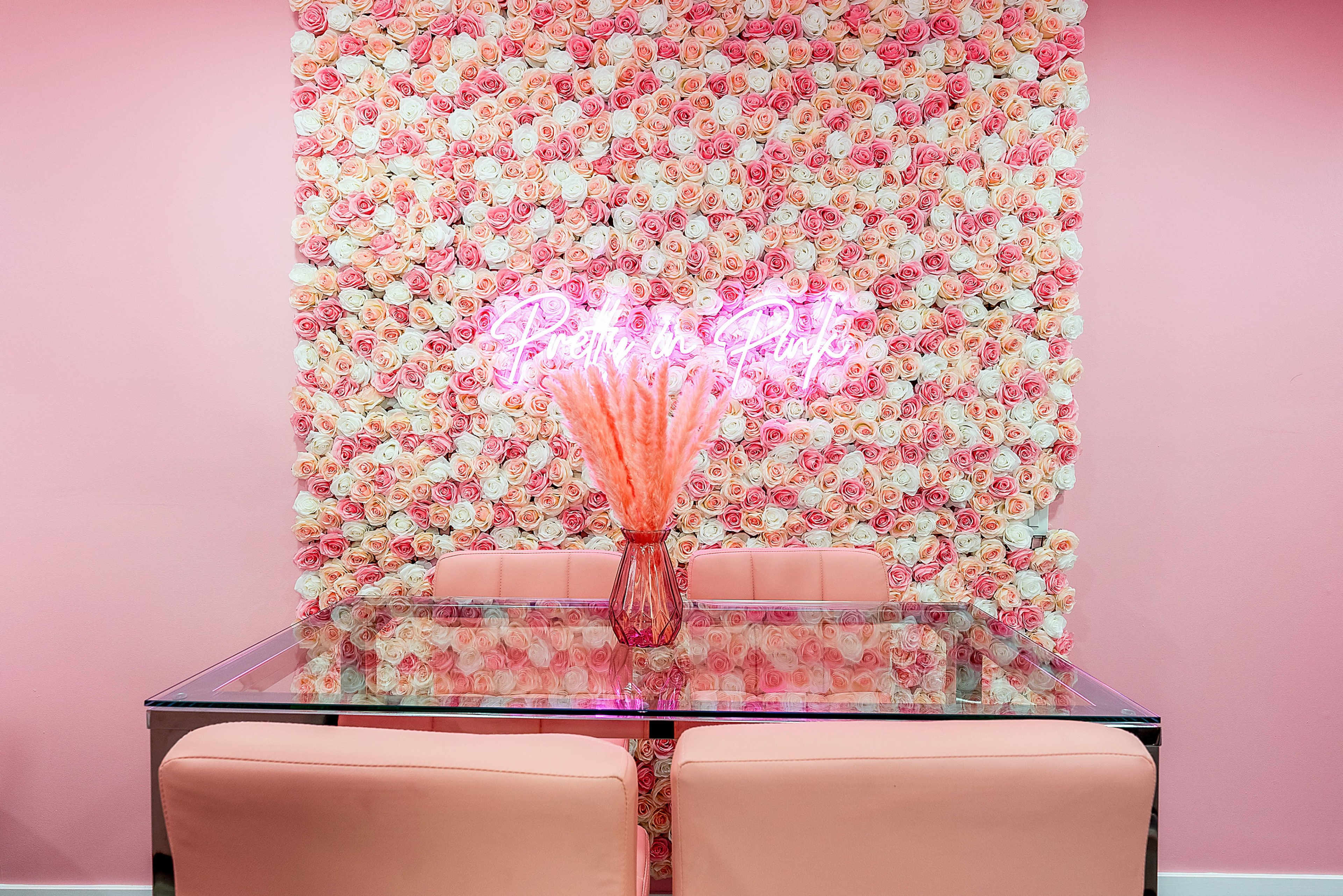 A glass table with pink chairs is placed in front of a wall covered in pink and white roses, with a neon sign above it.