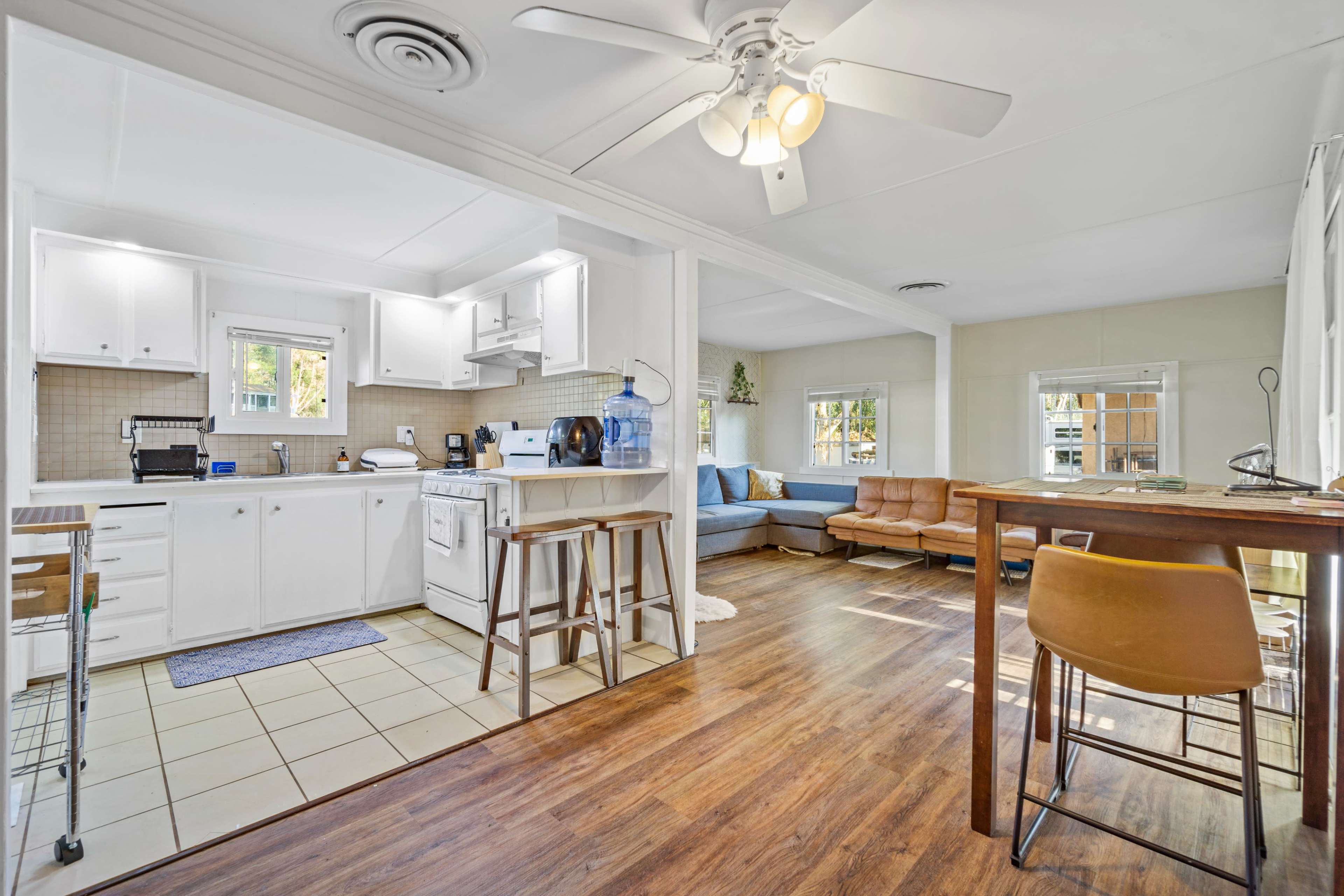 A bright, open-concept living space featuring a kitchen with white cabinets, a dining area with wooden seating, and a comfortable living room with a sofa.