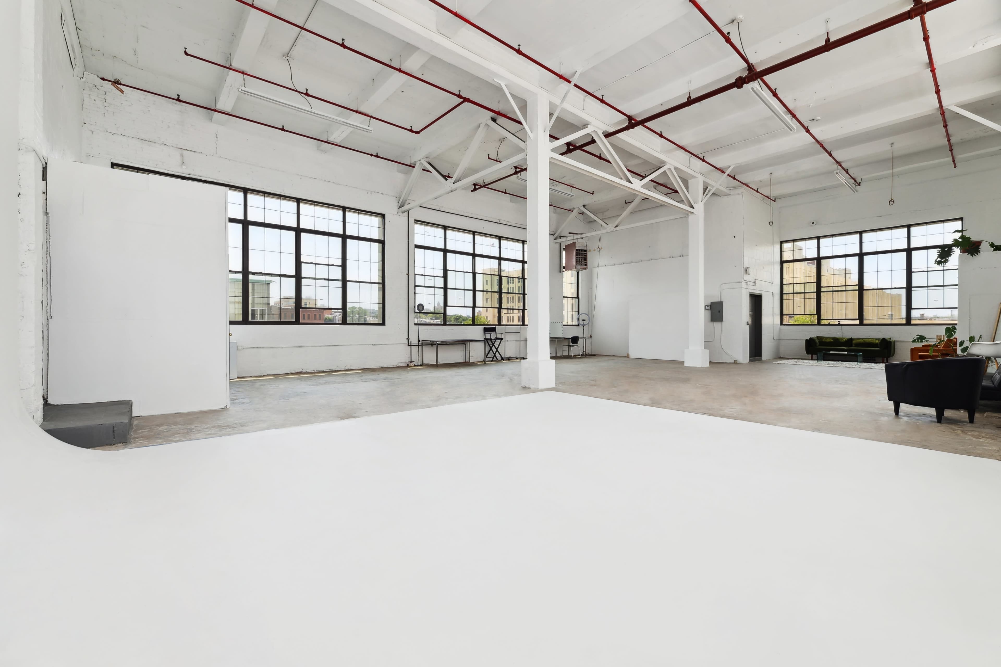 A spacious, sunny studio with large windows, a blank white wall, and minimal furniture.