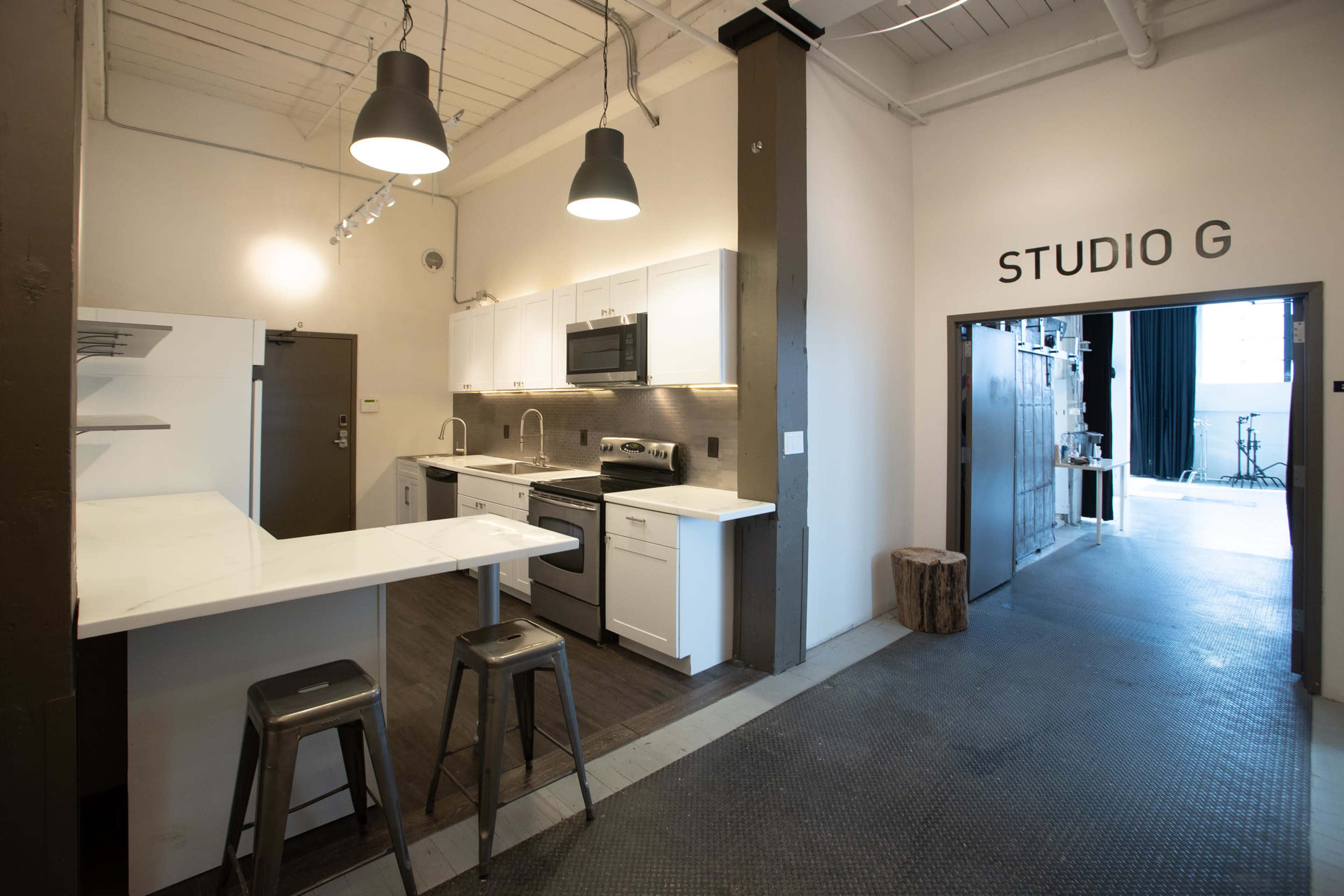 A modern kitchen area adjacent to an entrance labeled "STUDIO G," featuring white cabinetry, stainless steel appliances, and bar seating.