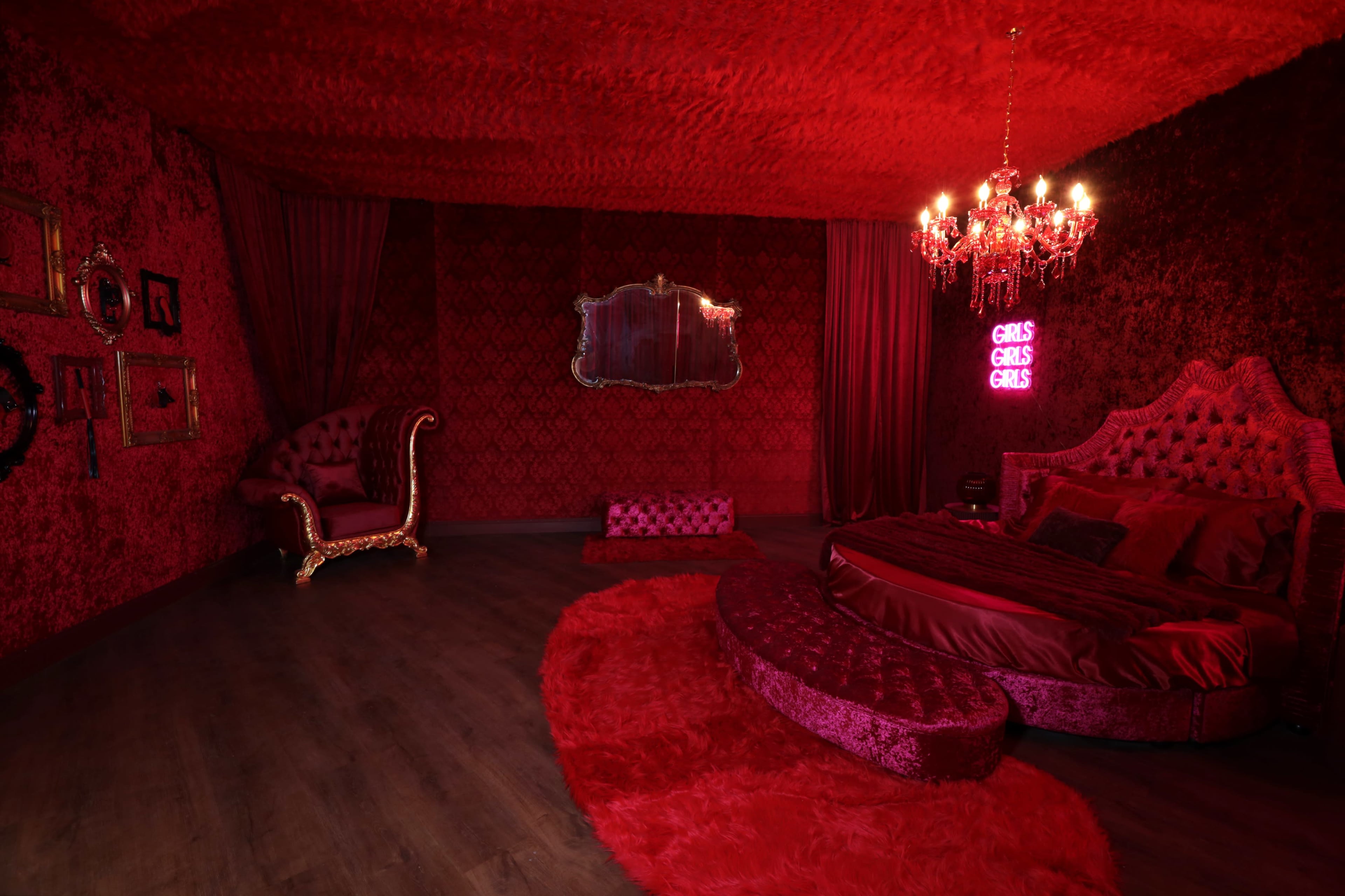 The room features red velvet walls, a plush bed, a pink rug, and a chandelier, with neon sign that reads "GIRLS GIRLS GIRLS."