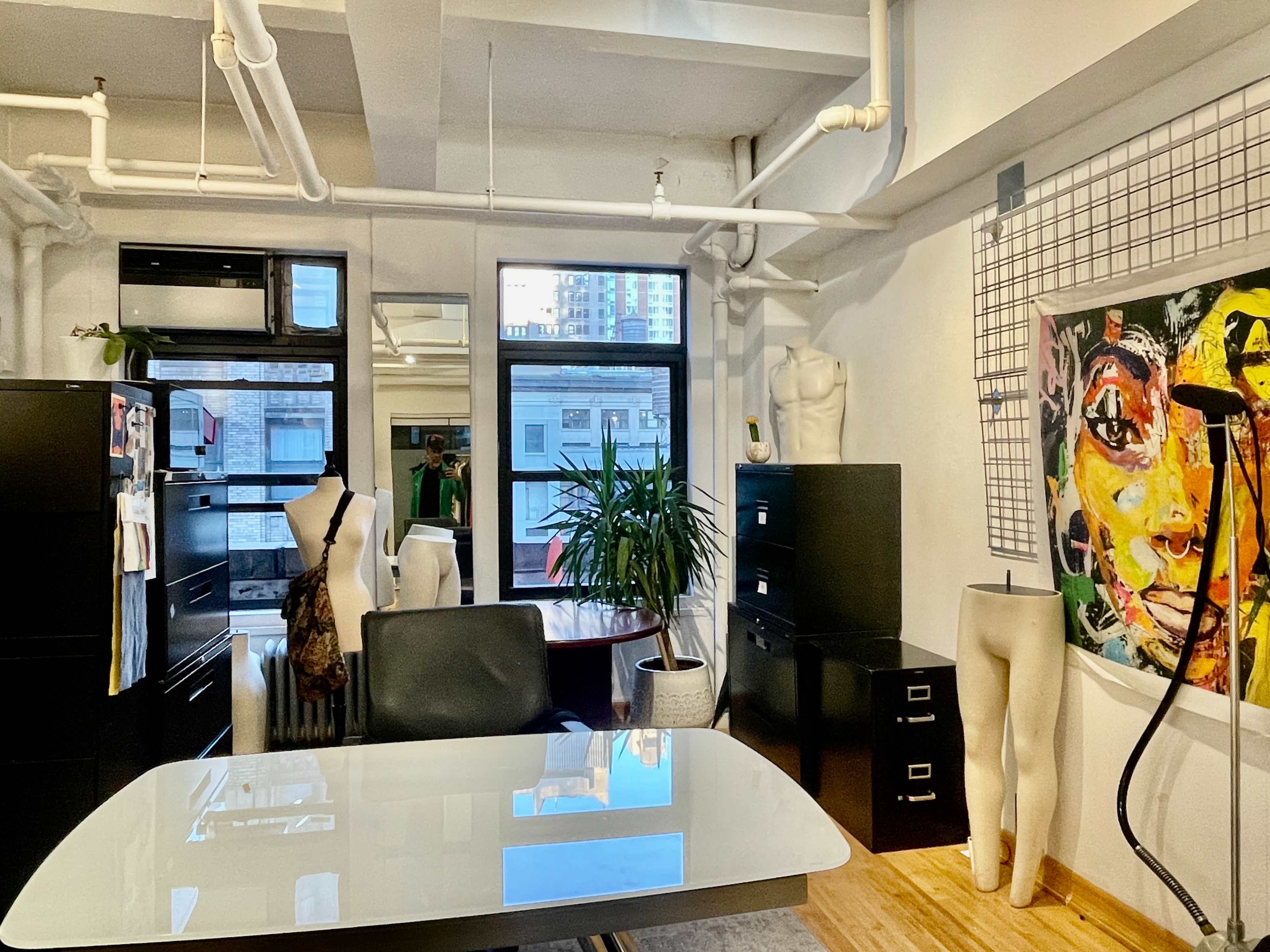 The image shows a modern office space featuring a glass table, black filing cabinets, a plant, and a wall with art and mannequin displays.