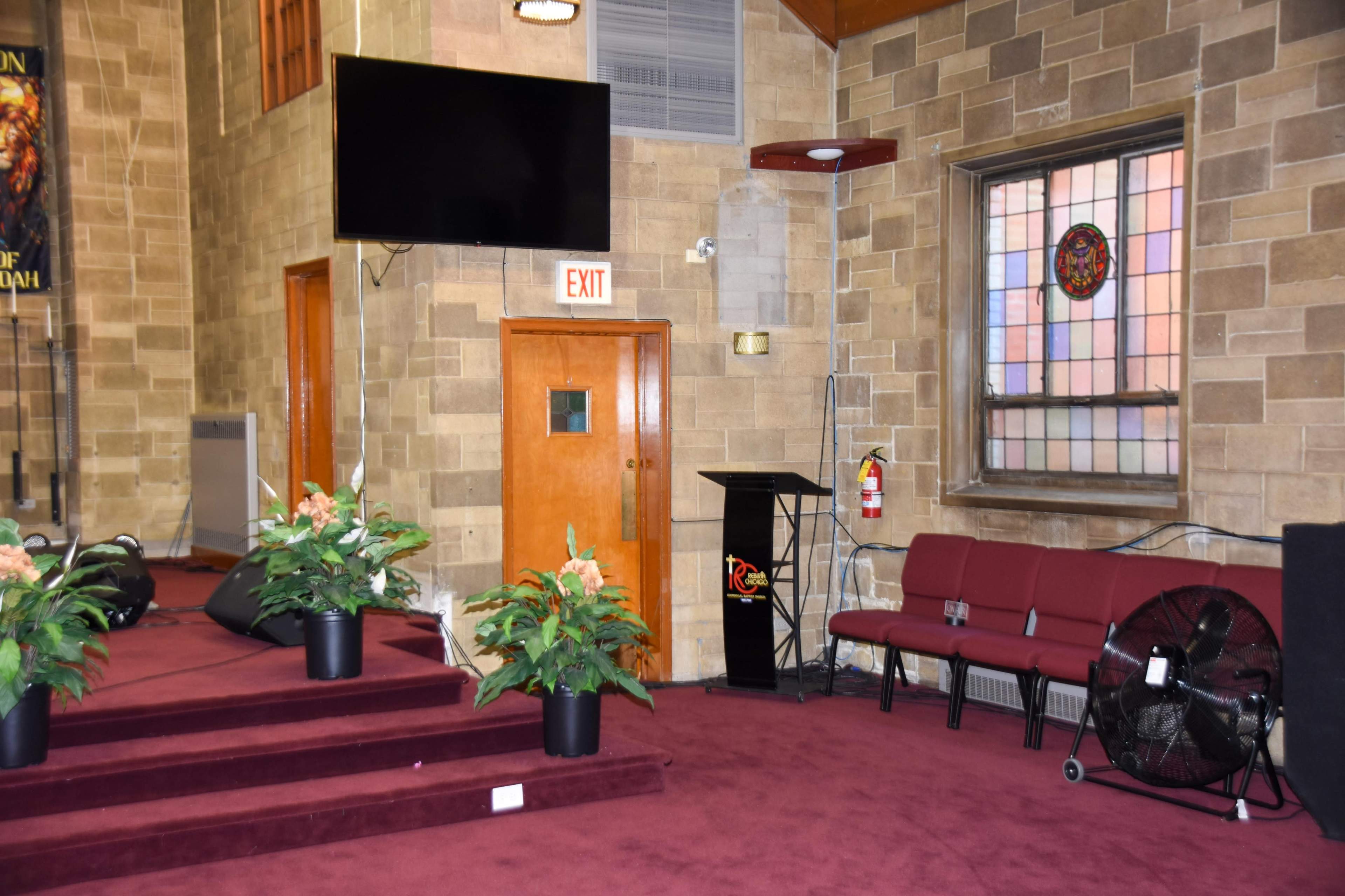 The scene features a small chapel or church interior with stone walls, a raised platform, red carpeting, potted plants, and a few chairs arranged near a podium.