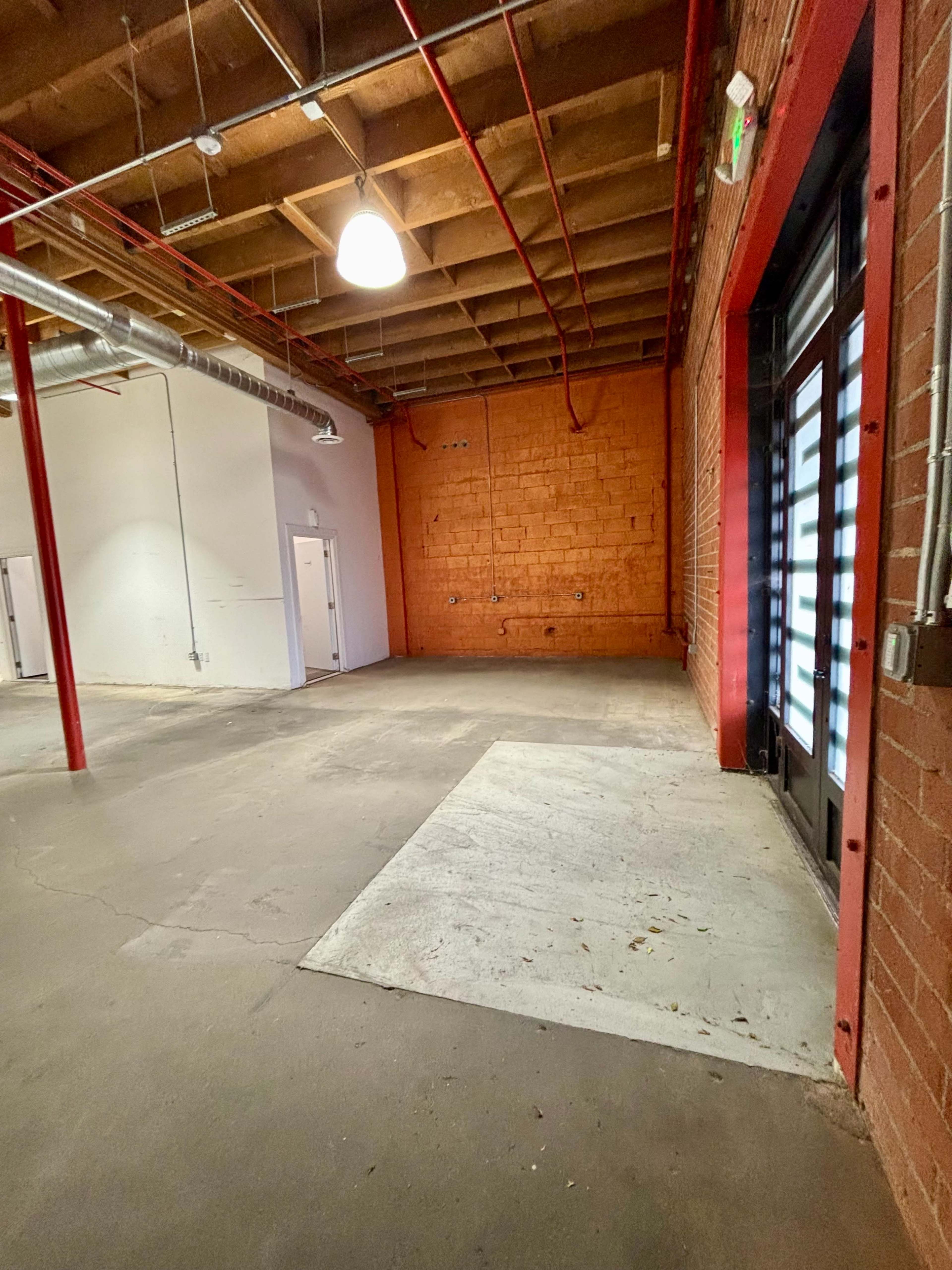 The image shows an empty industrial space with exposed red brick walls, concrete flooring, and overhead piping.
