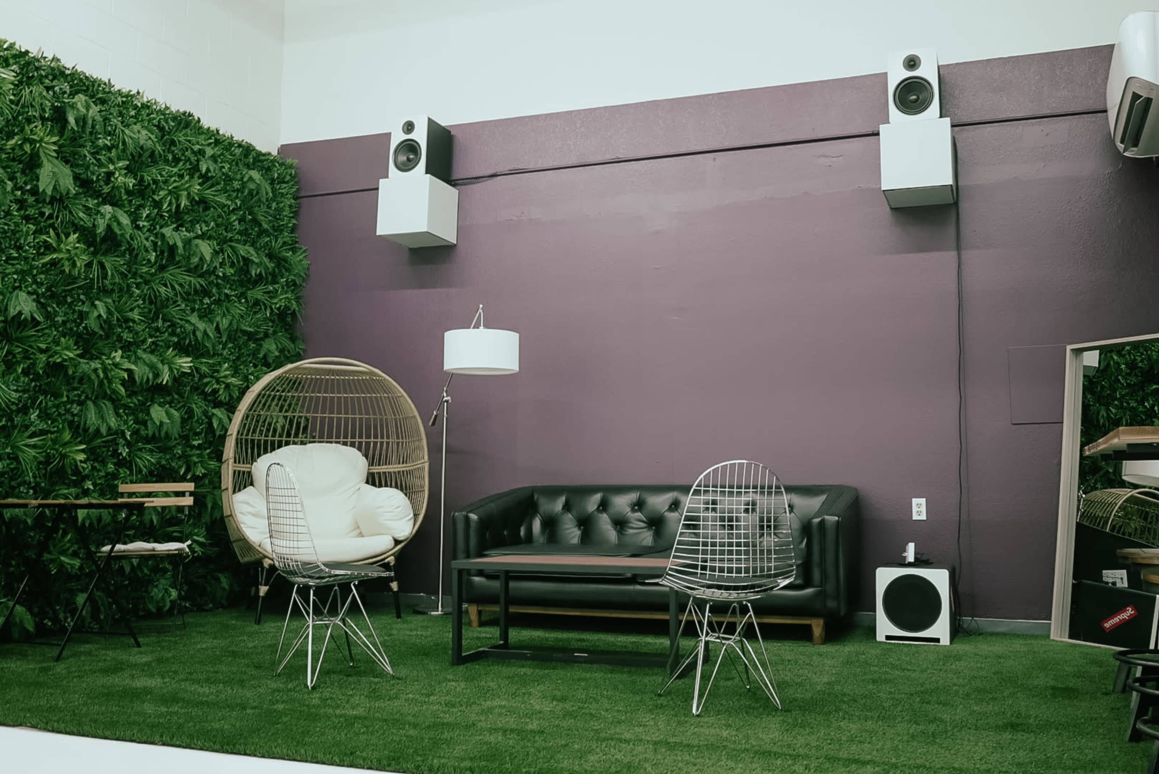 A modern indoor space featuring a green wall, a black leather sofa, and two wire chairs on a grassy carpet.