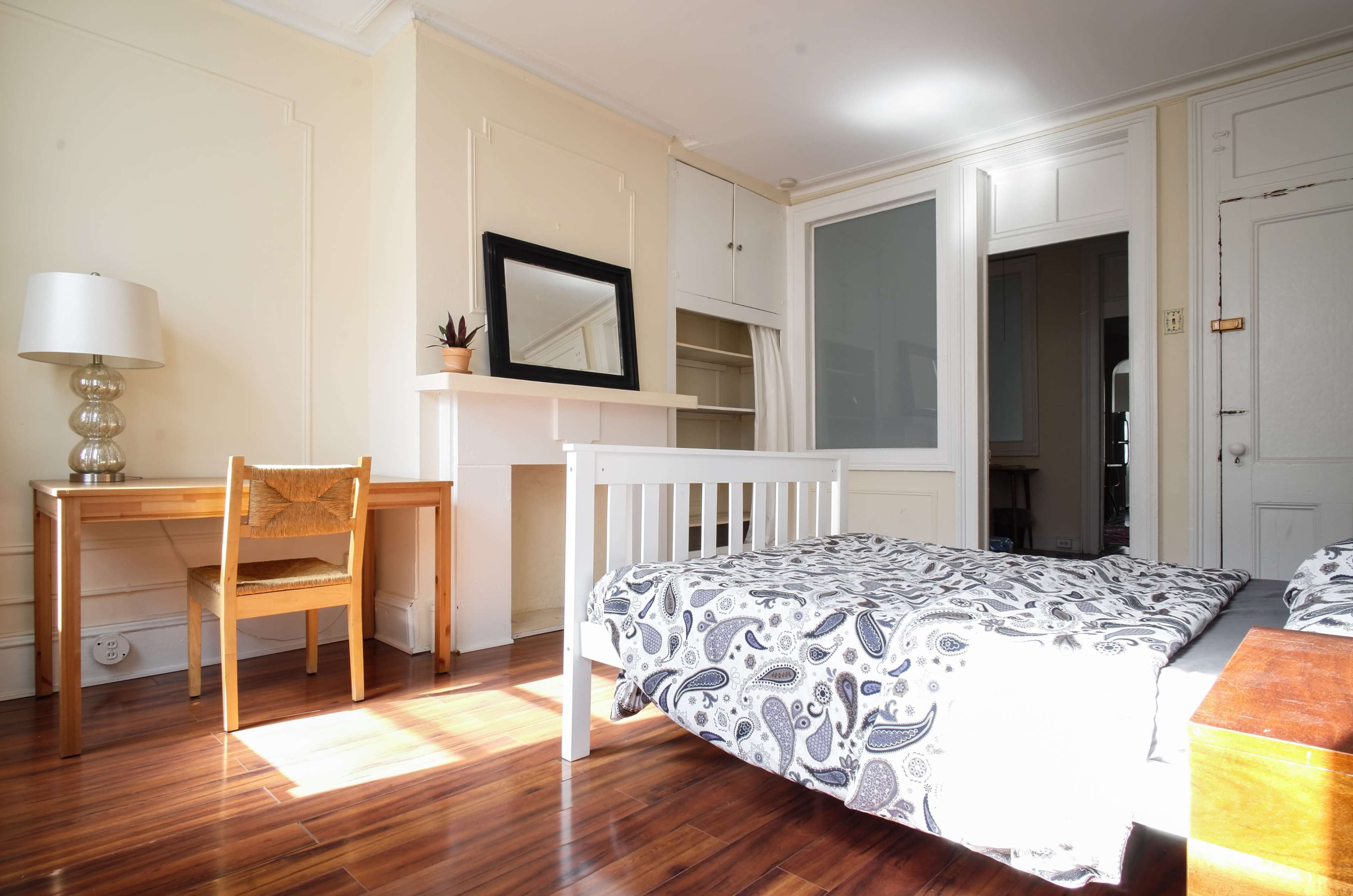 A small, bright bedroom featuring a bed with a patterned comforter, a wooden desk and chair, and a mirror on the wall above a fireplace.