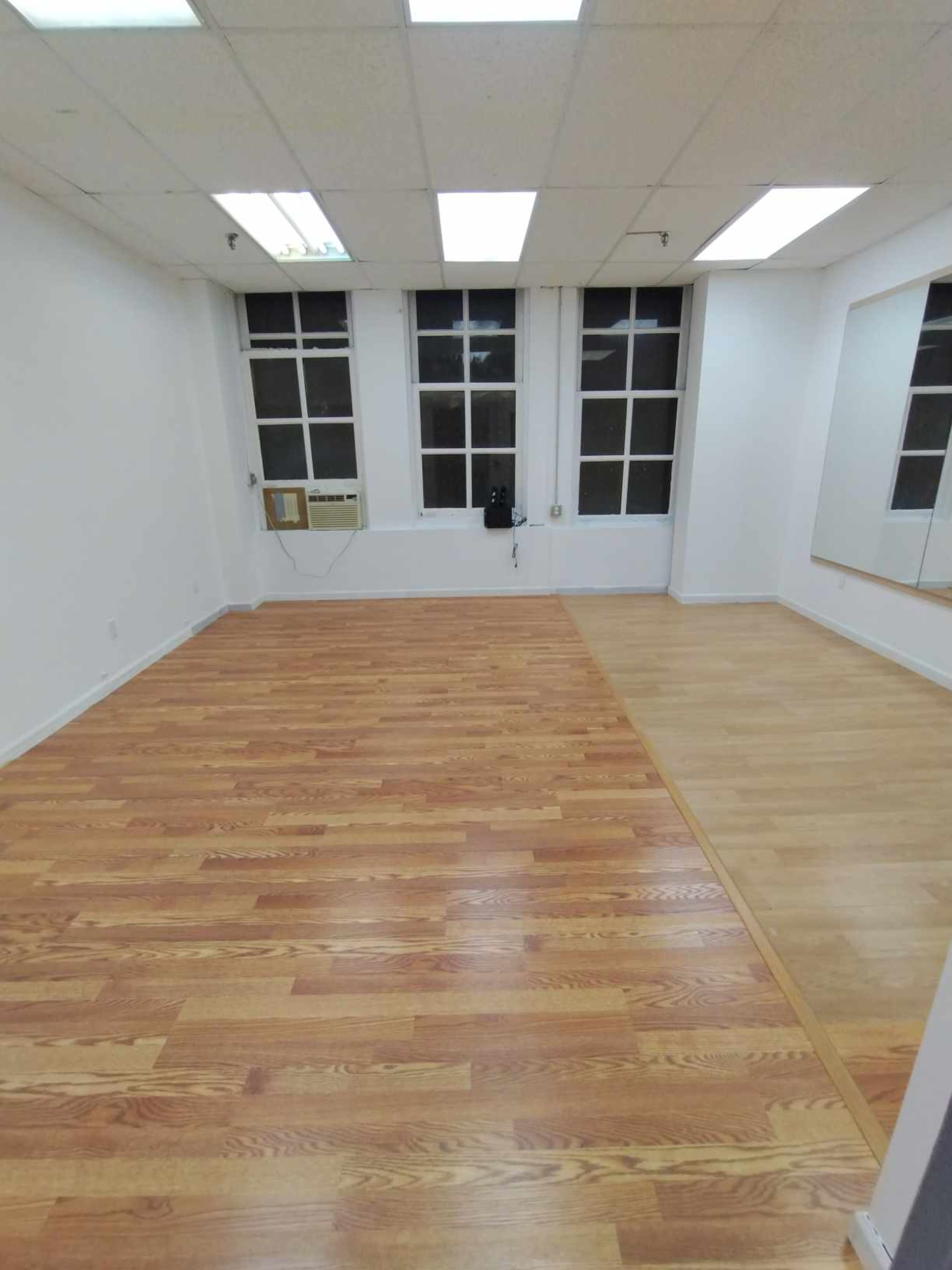 The image shows an empty room with hardwood flooring, large windows, and a mirror on one wall.