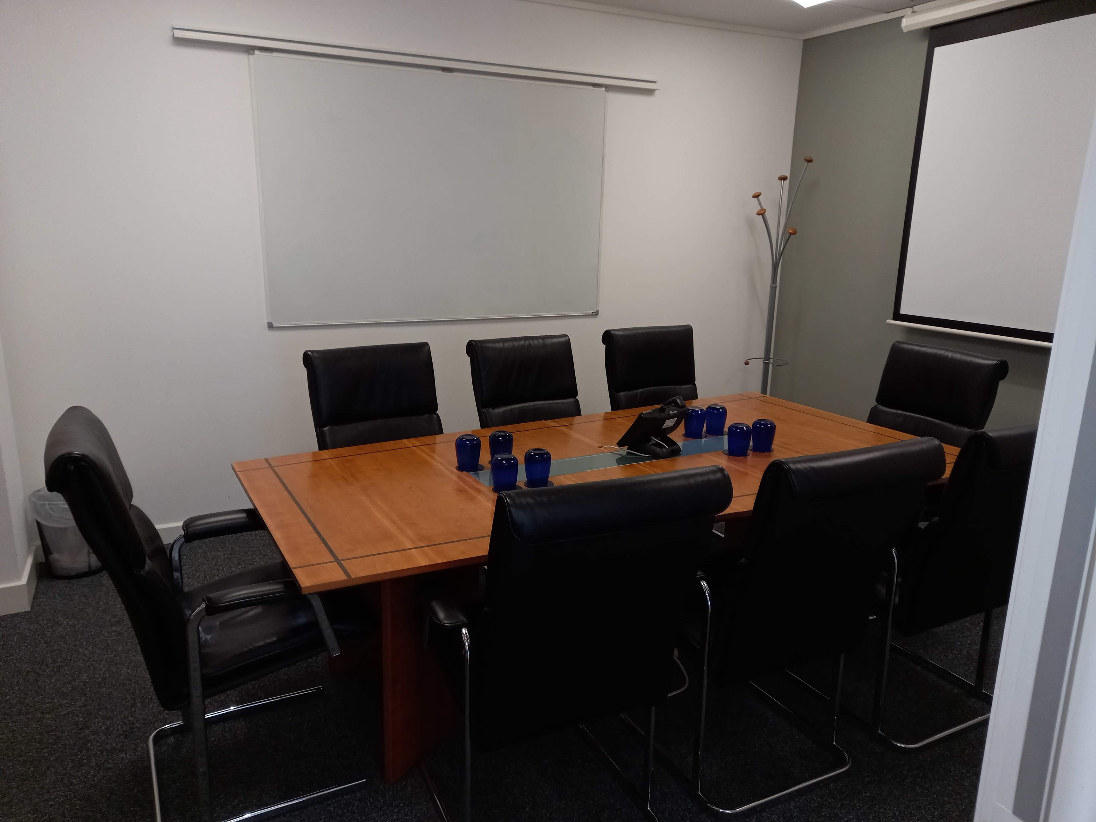 A conference room features a large wooden table surrounded by black leather chairs, with a whiteboard and projector screen on the wall.