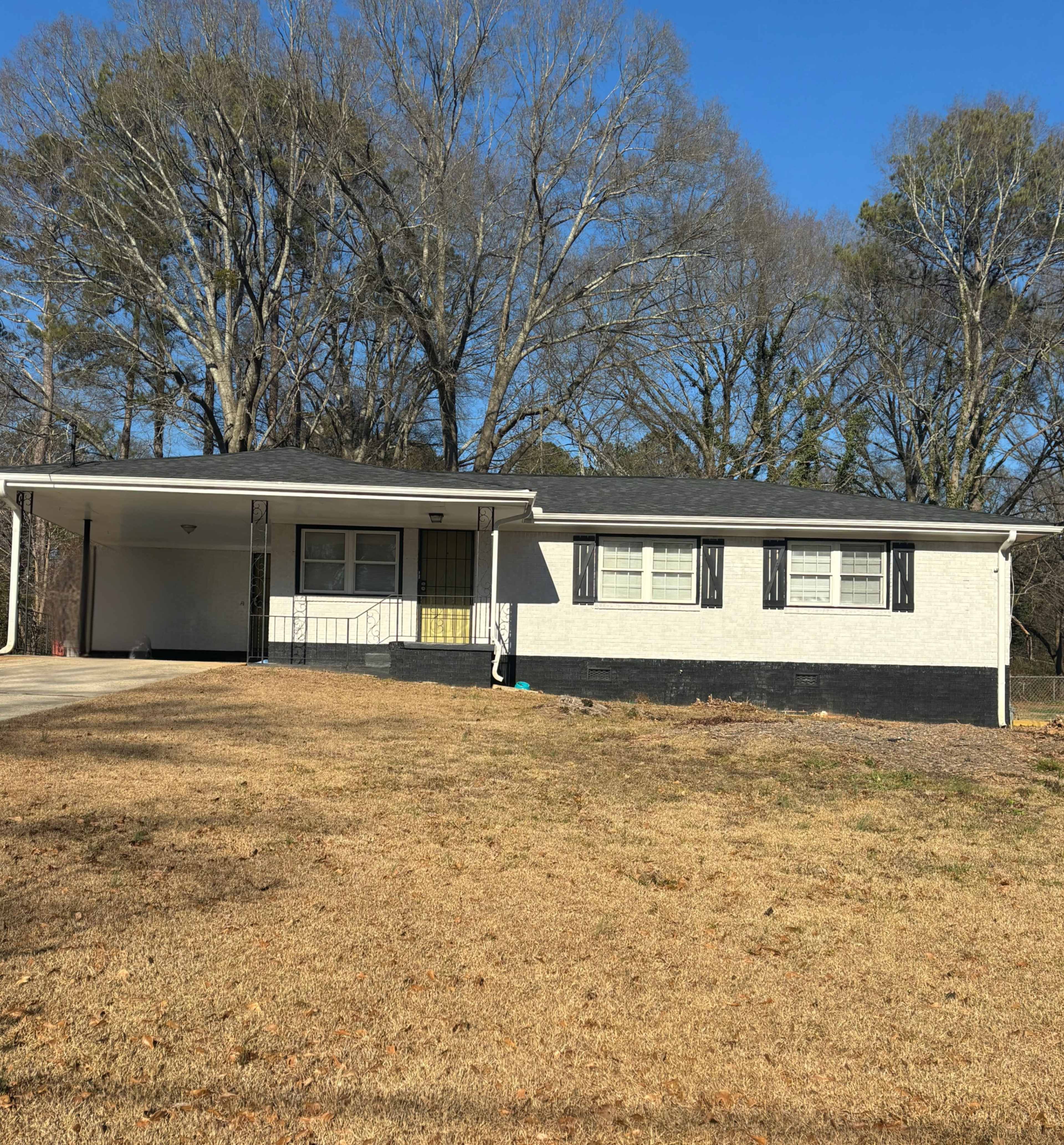 A single-story house with a carport, white brick exterior, and large windows is situated on a grassy lawn surrounded by trees.