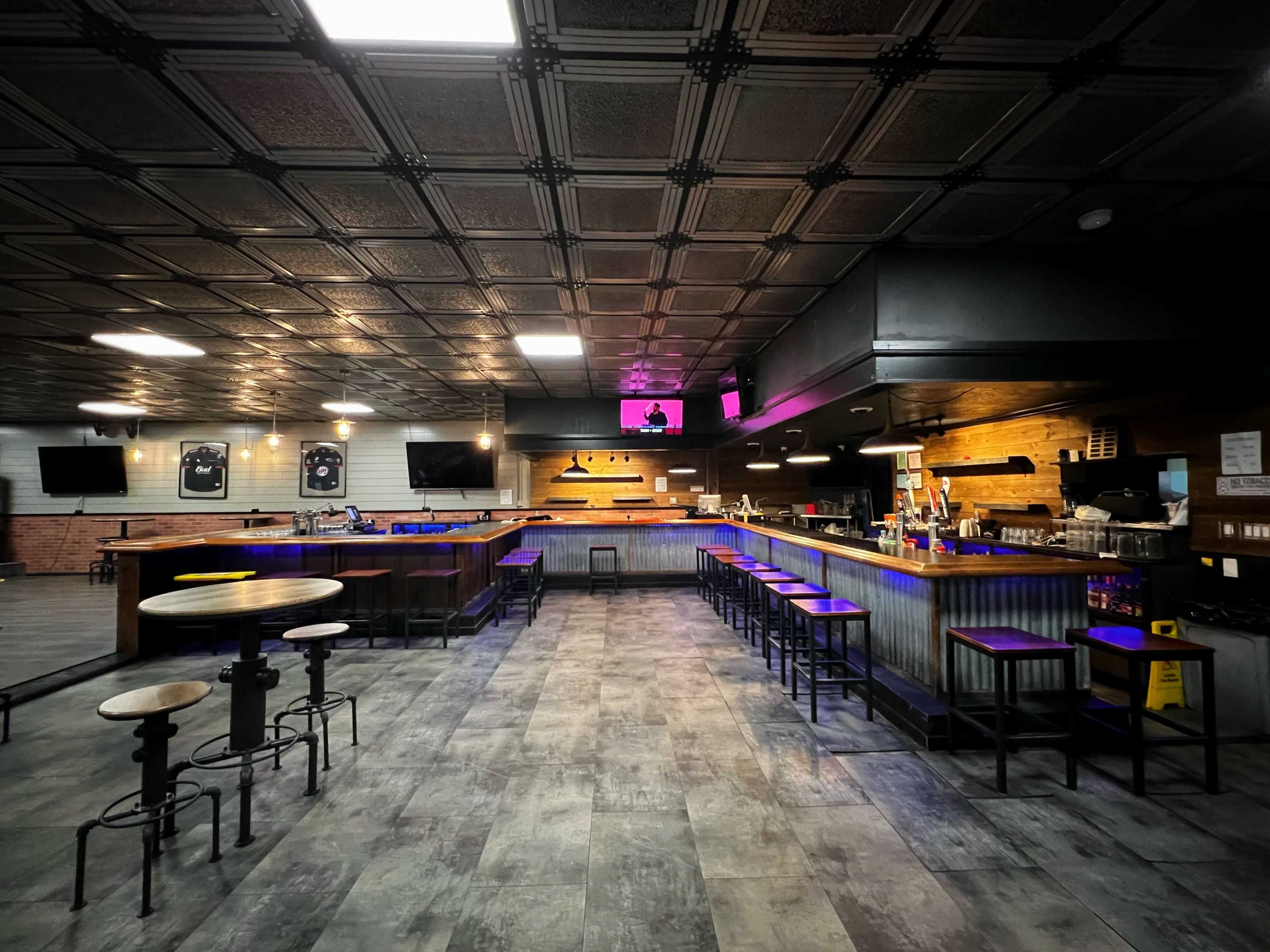 The interior of a modern bar with a long counter, high stools, and several tables arranged across the floor.