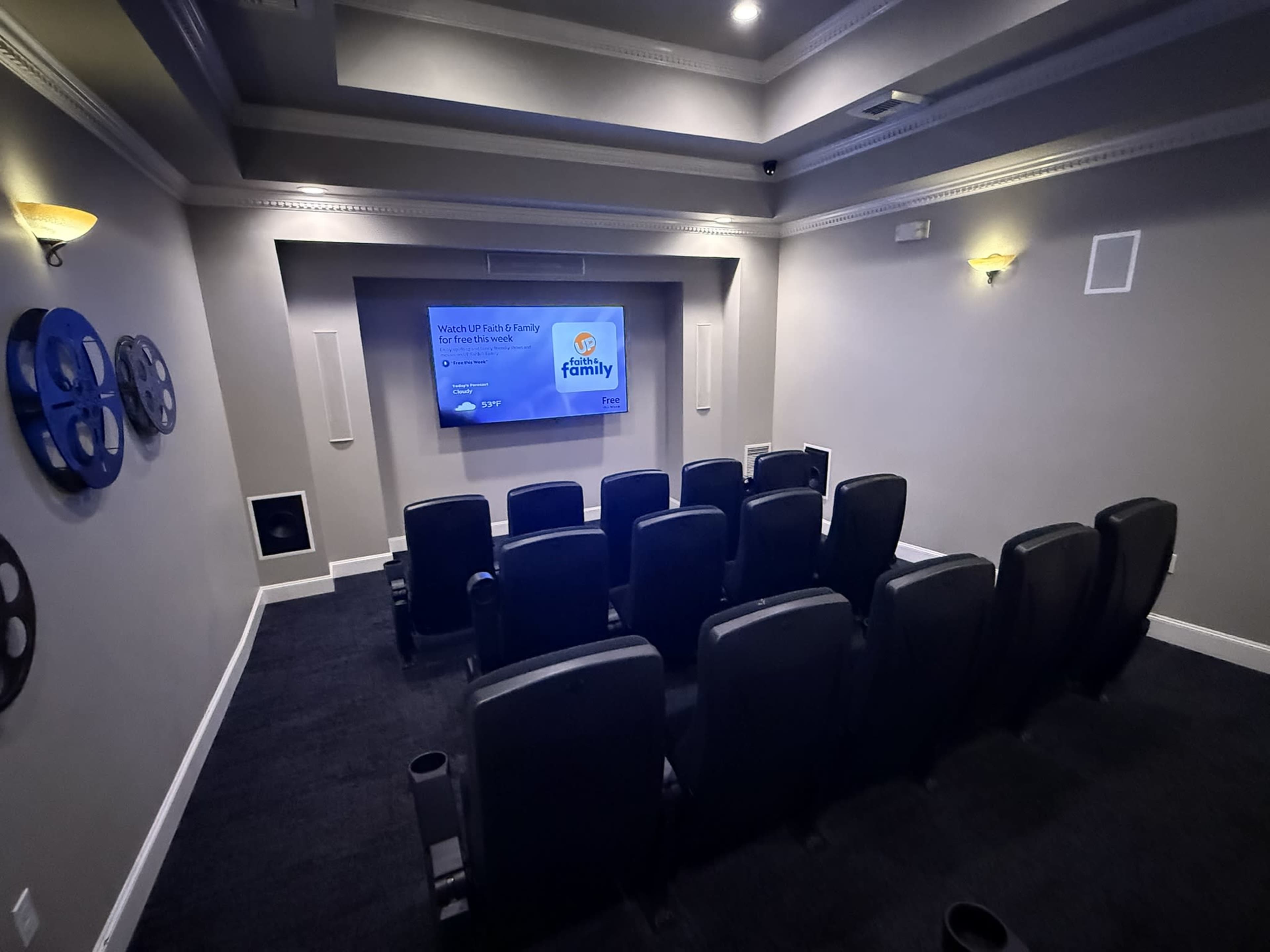 The image shows a small, dark home theater with black seating arranged in rows facing a large screen displaying a logo.