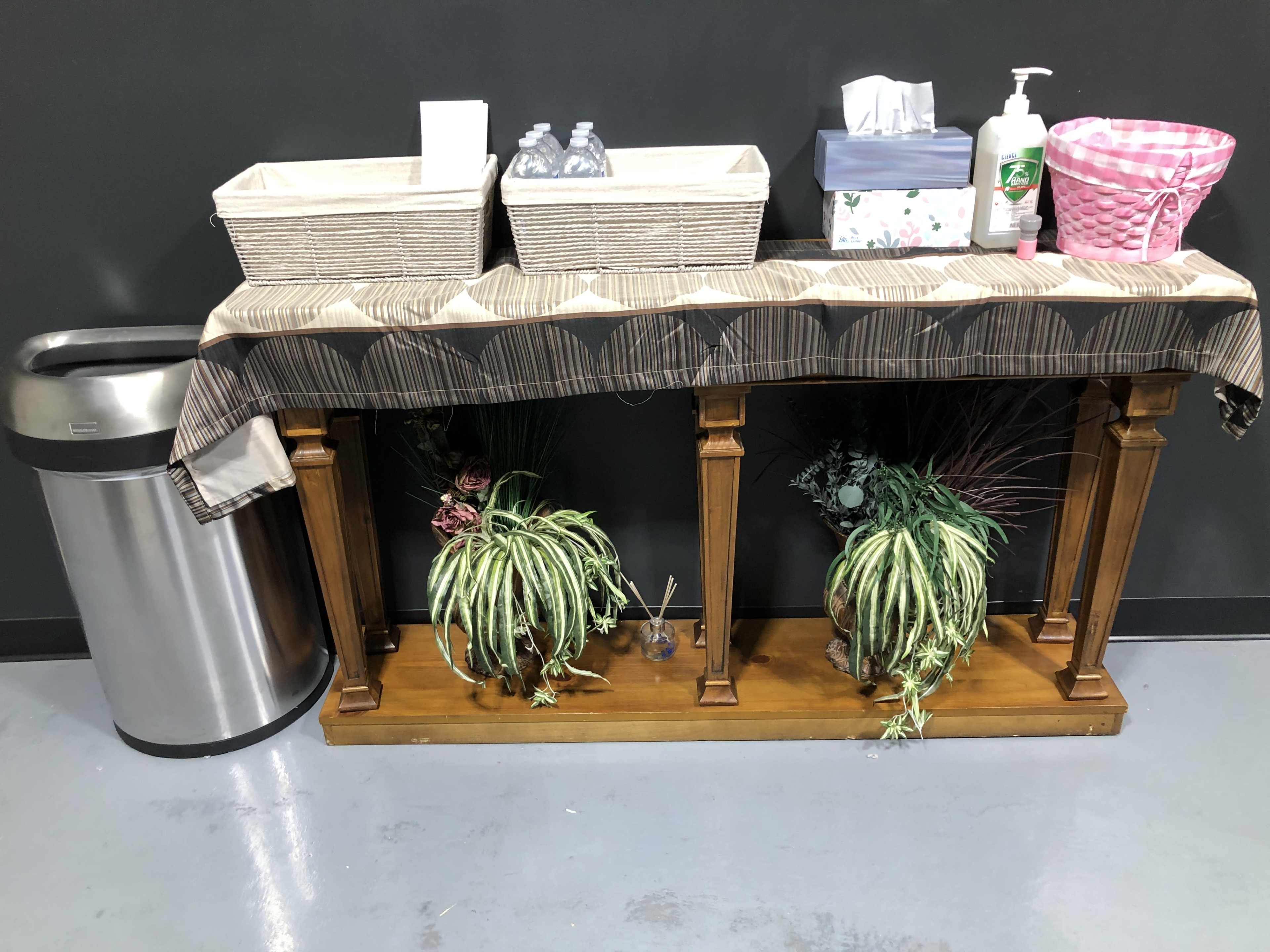 A wooden table with a patterned cloth displays two baskets, hand sanitizer, a box, and two potted plants, positioned beside a trash bin against a dark wall.