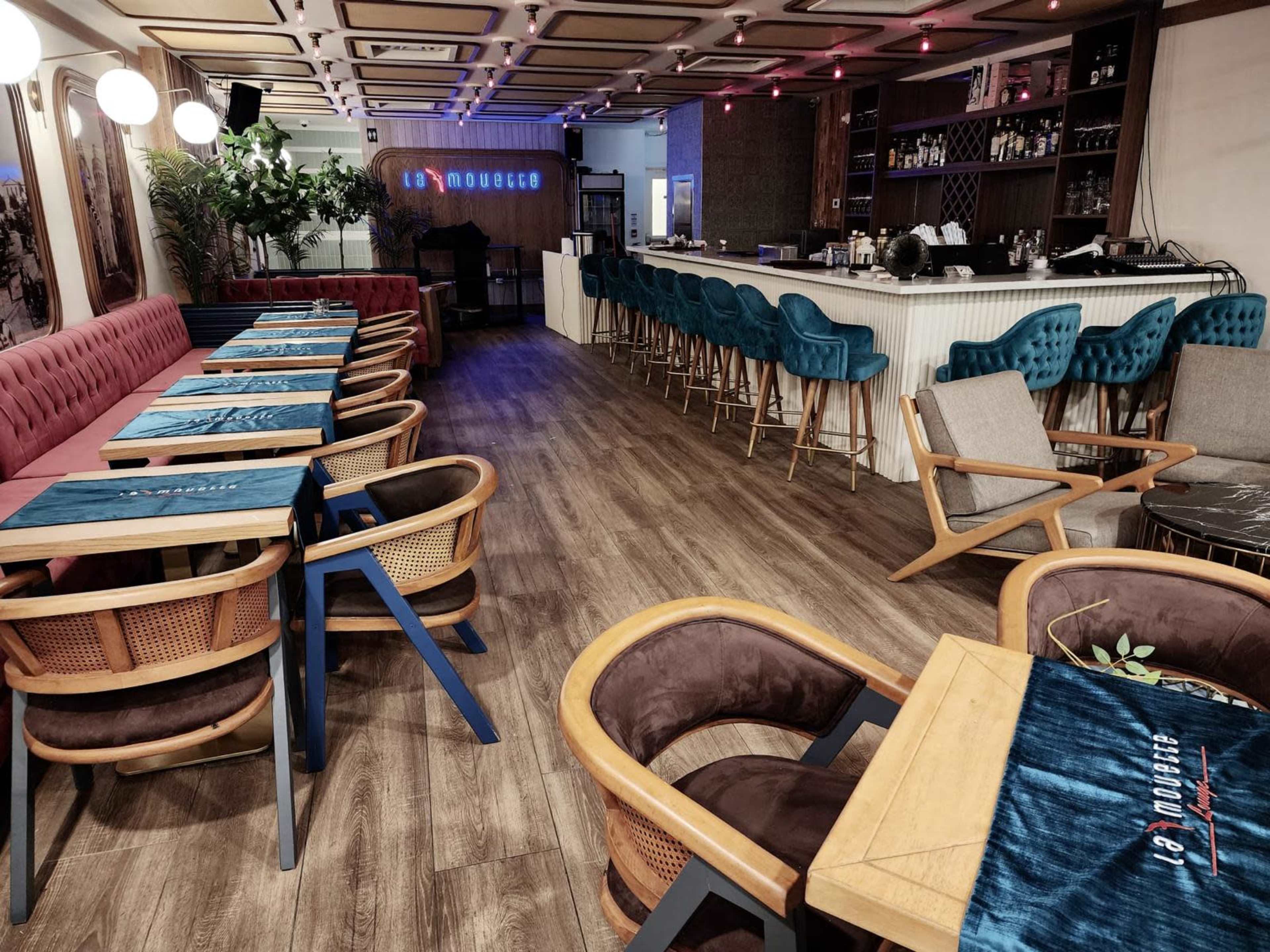 The image shows a modern cafe interior with wooden flooring, colorful seating, and a bar area in the background.