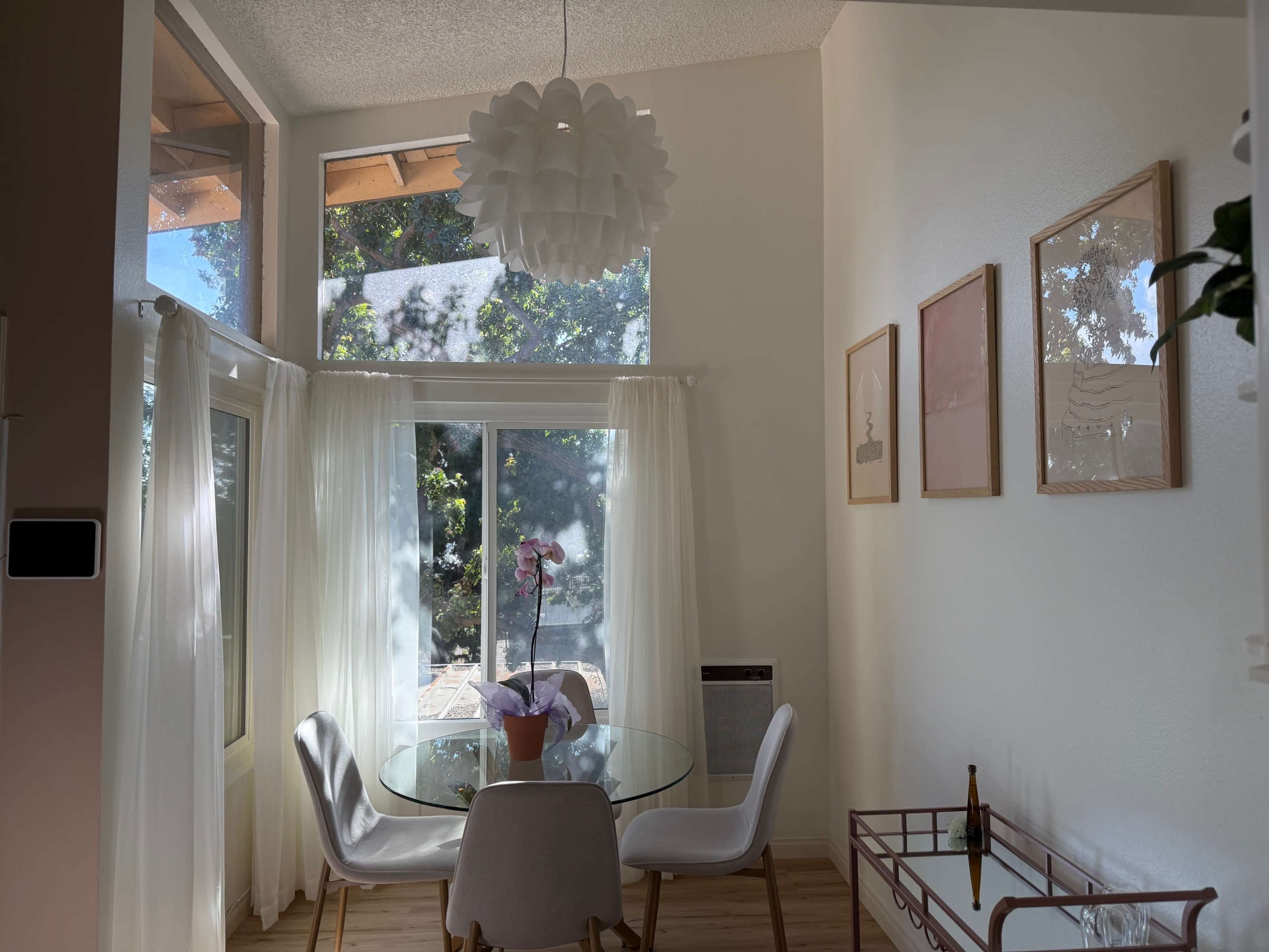 A dining area features a glass table surrounded by white chairs, with a large window providing natural light and a view of greenery outside.
