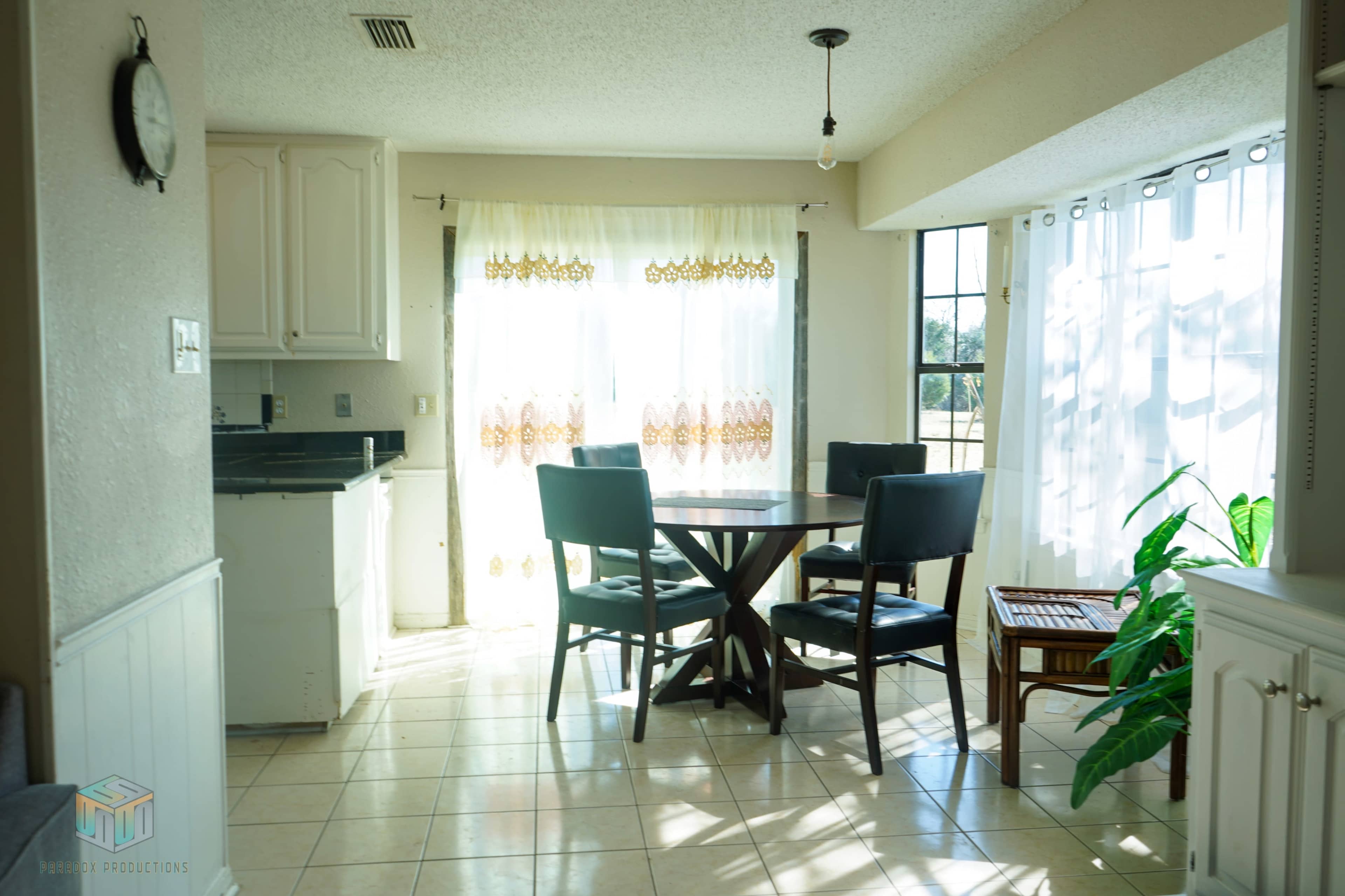 A bright dining area features a round table surrounded by four black chairs, with large windows dressed in sheer curtains allowing natural light to flood the space.