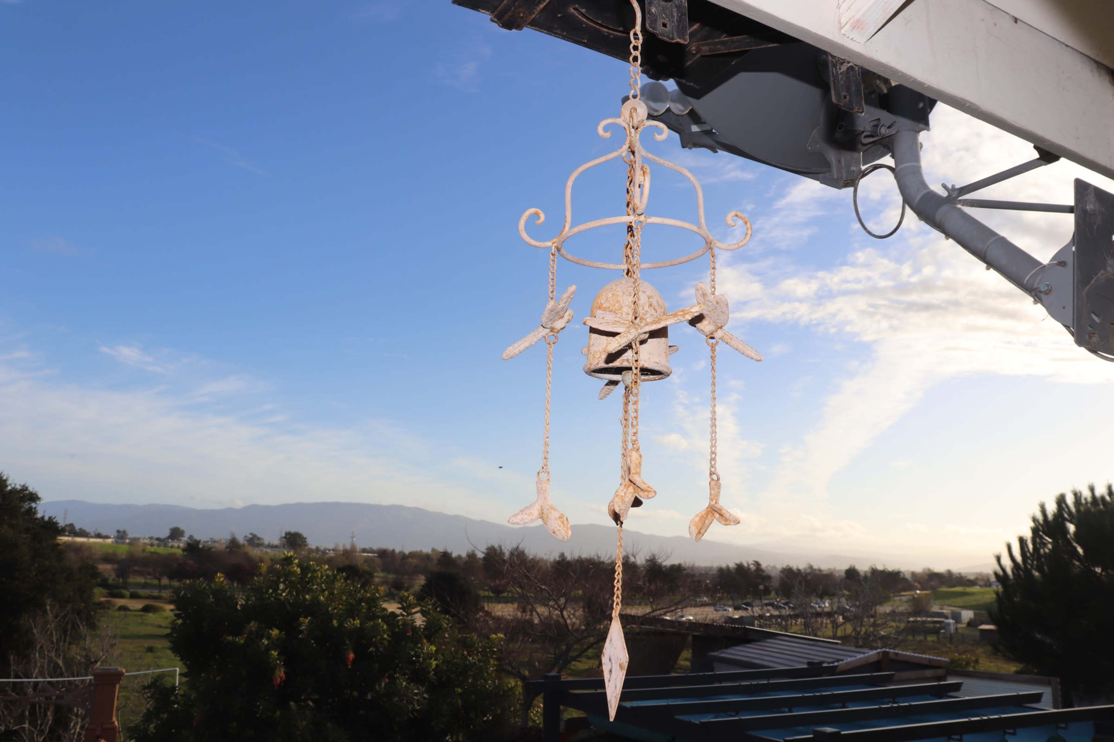 A decorative wind chime hangs from an overhanging structure against a backdrop of rolling hills and a clear blue sky.