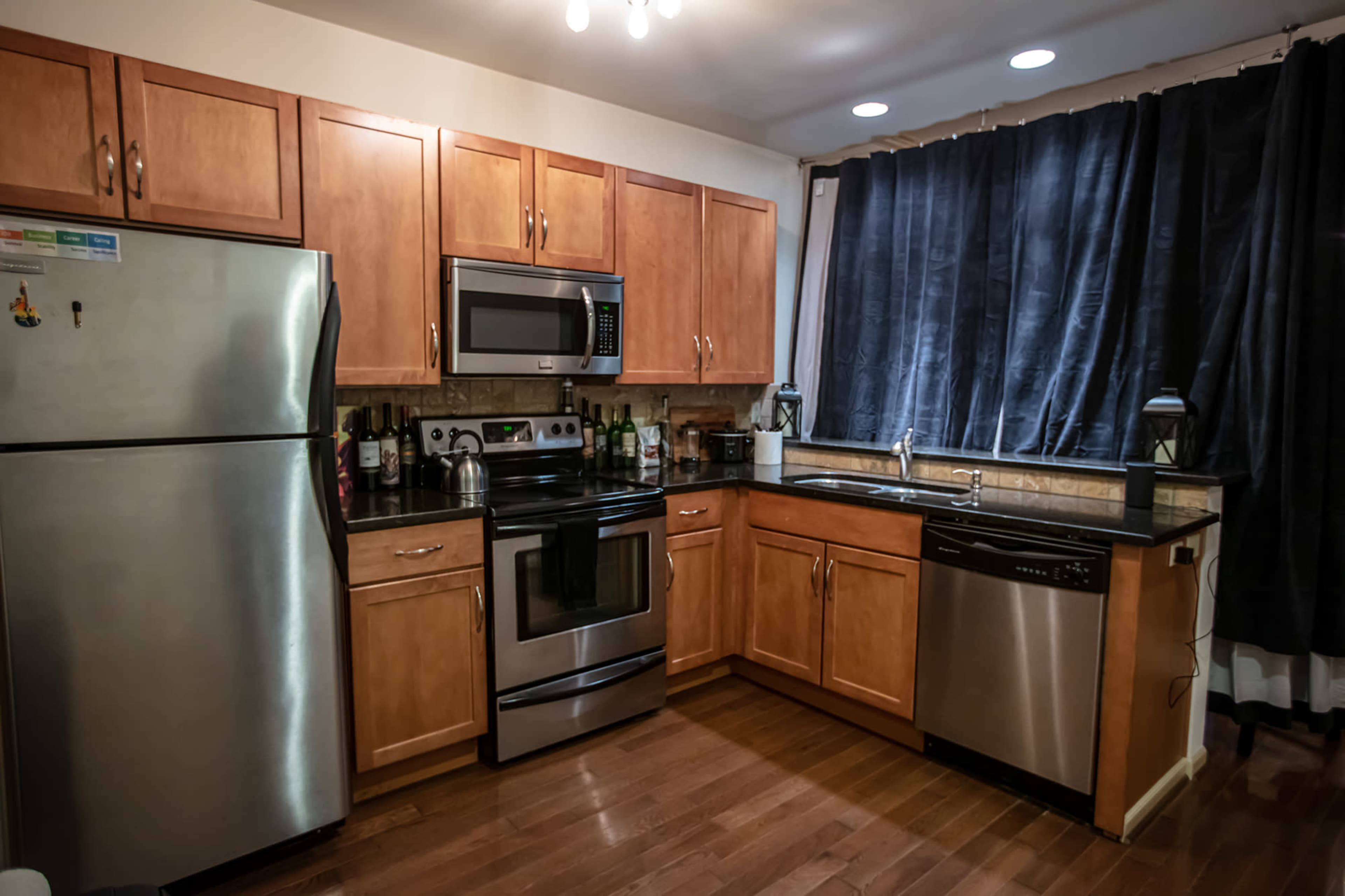 A kitchen equipped with wooden cabinets, a stainless steel refrigerator, an oven, a microwave, and a dishwasher, with dark curtains on the window.