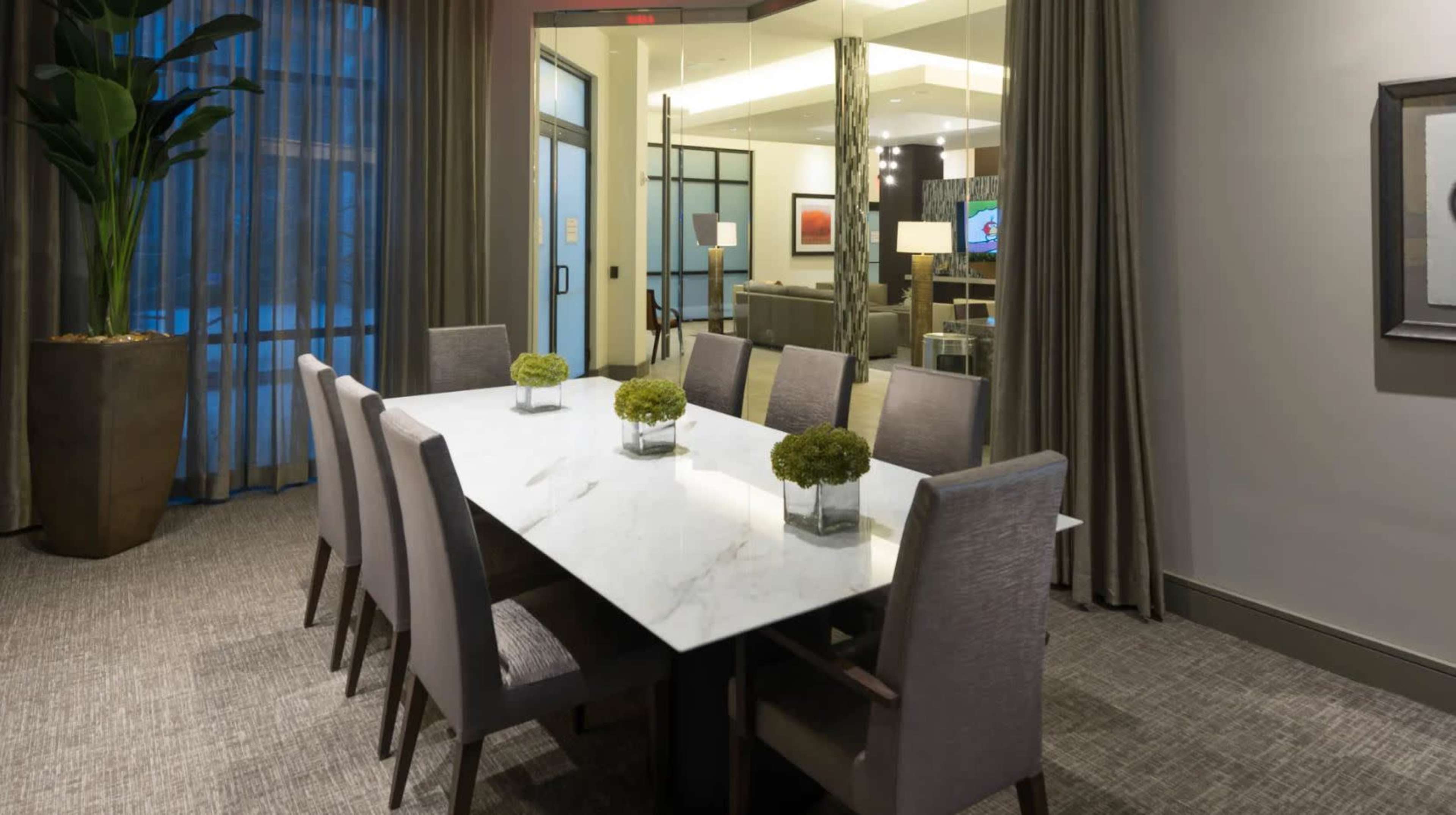 A modern conference room features a long marble table surrounded by six chairs, with potted greenery accents, and large windows draped with curtains.