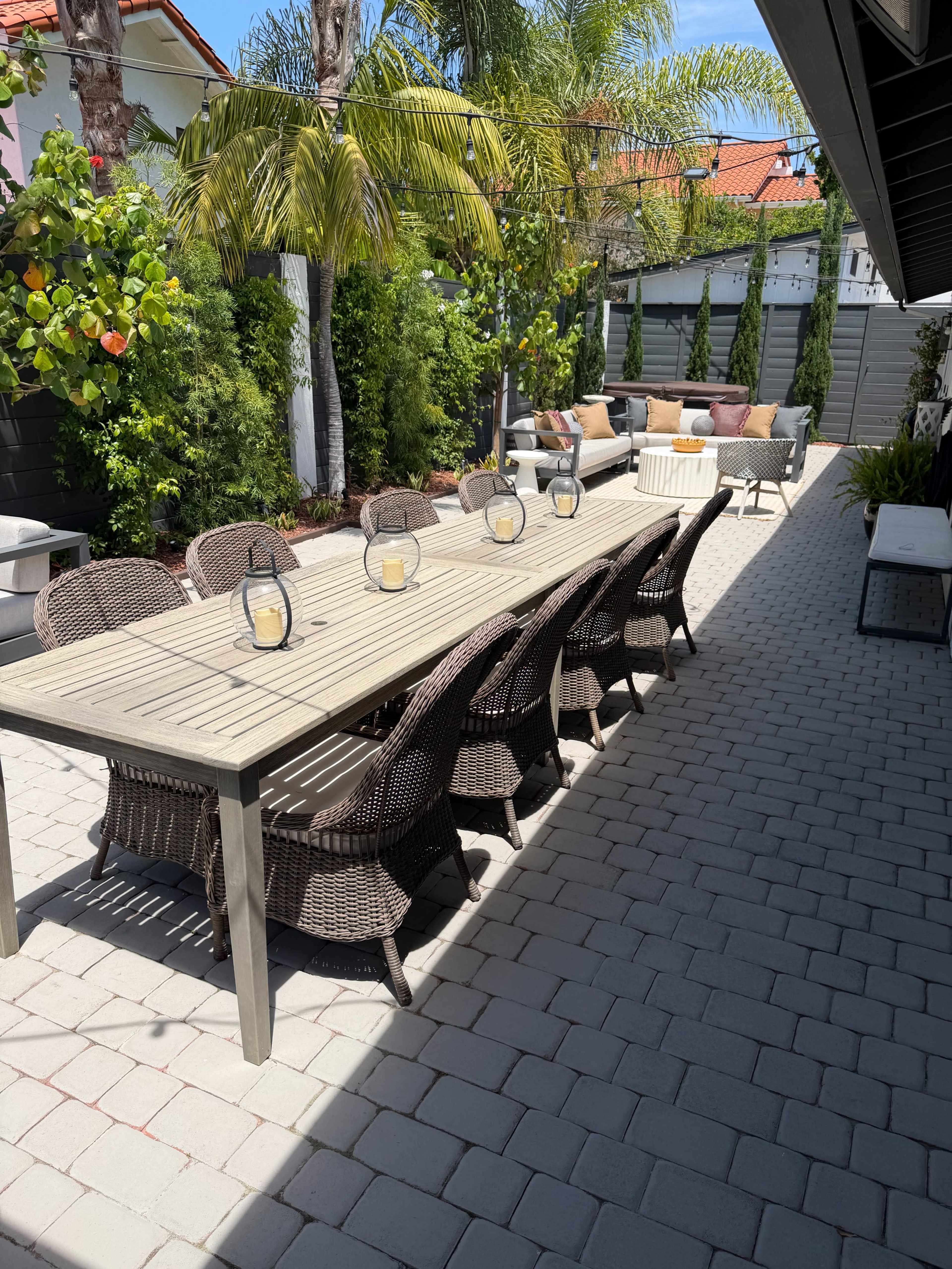 The image shows a long wooden dining table surrounded by wicker chairs in a sunlit patio with lush greenery and potted plants.