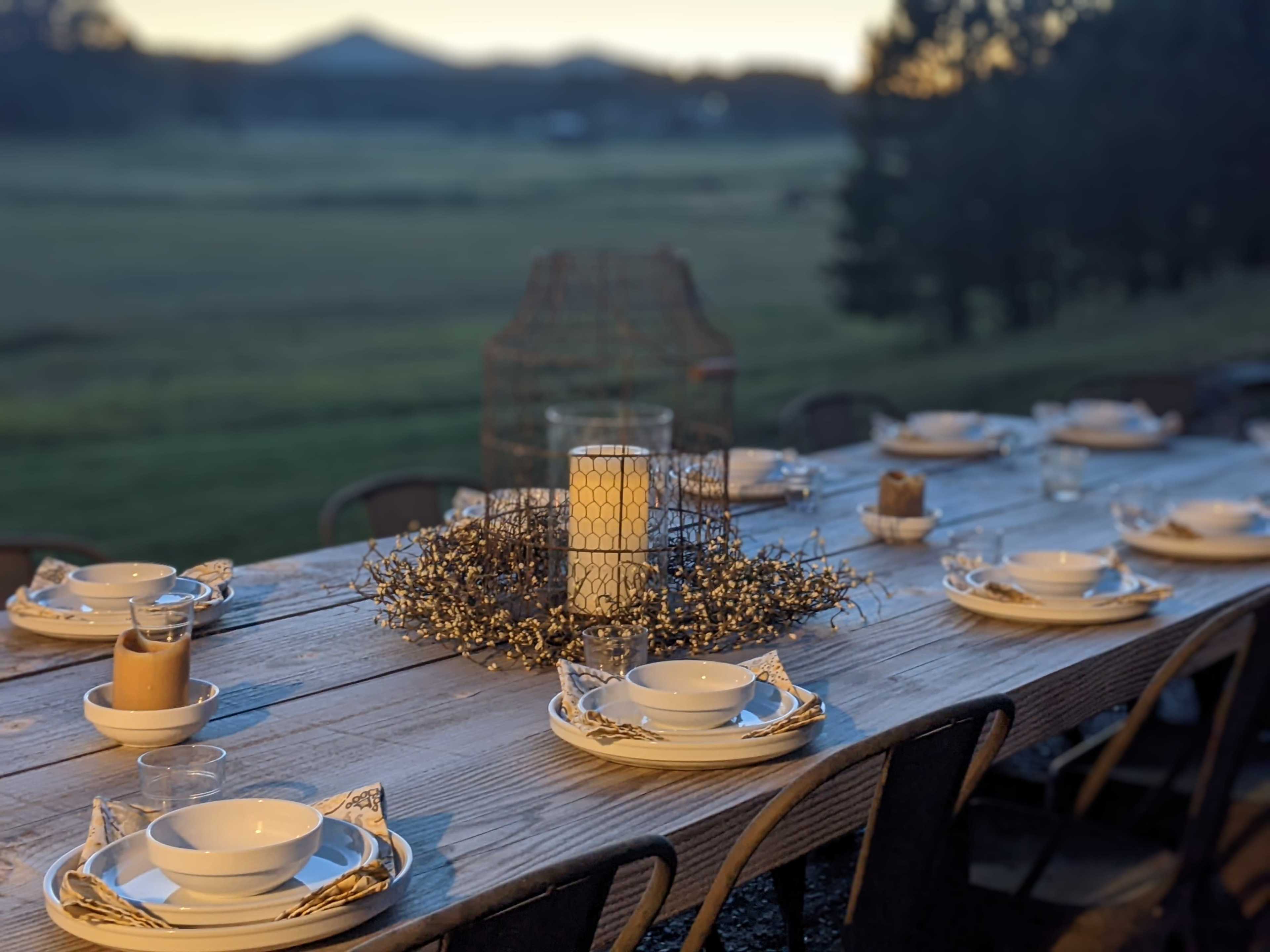 A long wooden table is set for a meal, featuring white plates, glasses, and decorative items, with mountains in the background and a sunset illuminating the scene.