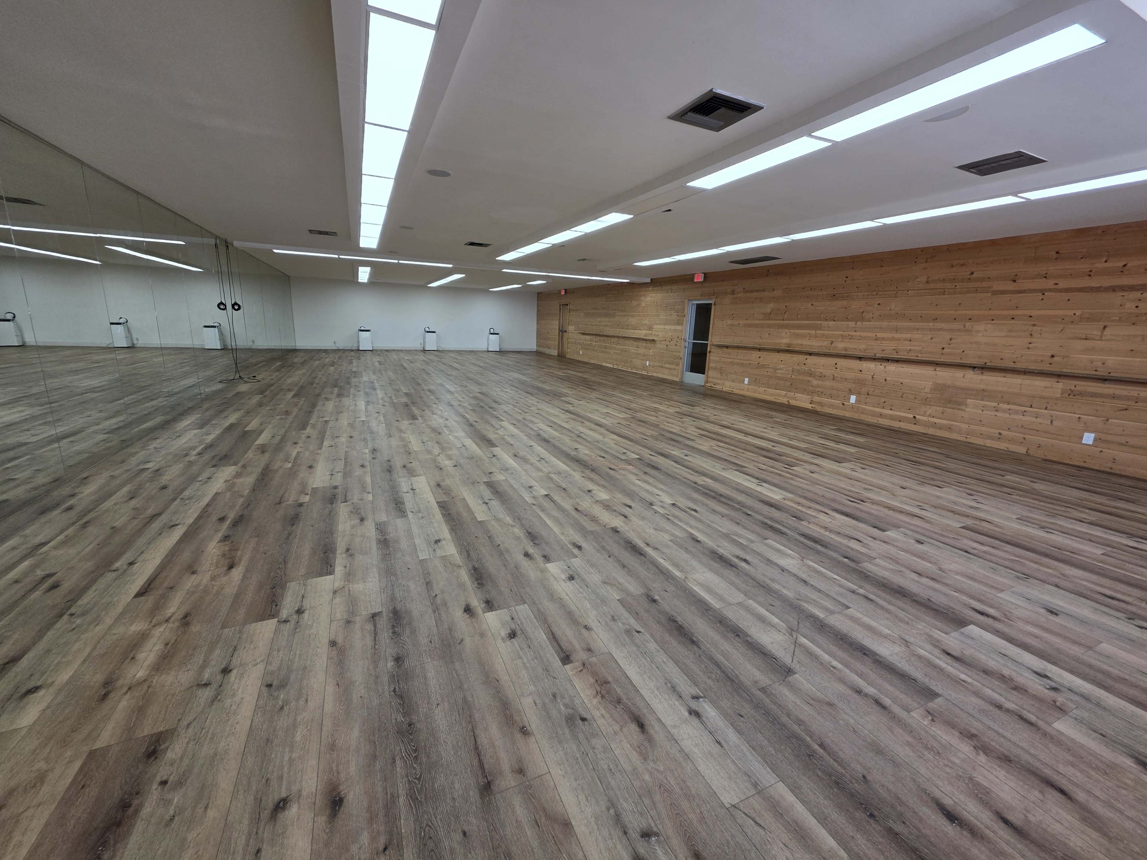The image shows a spacious, empty room with wooden flooring and mirrored walls.