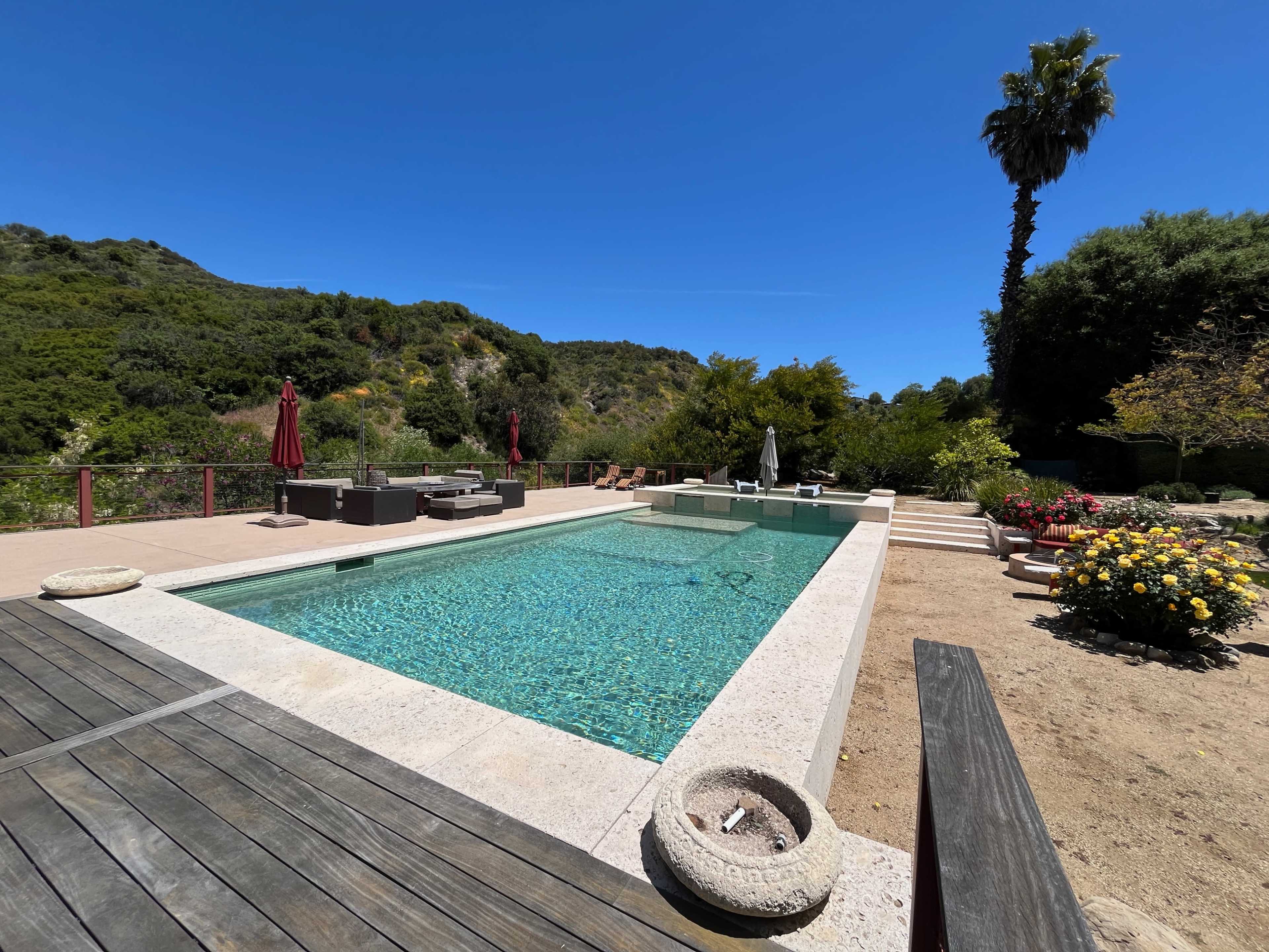 The image shows a long swimming pool surrounded by a landscaped garden with seating areas, palm trees, and colorful flowers under a clear blue sky.