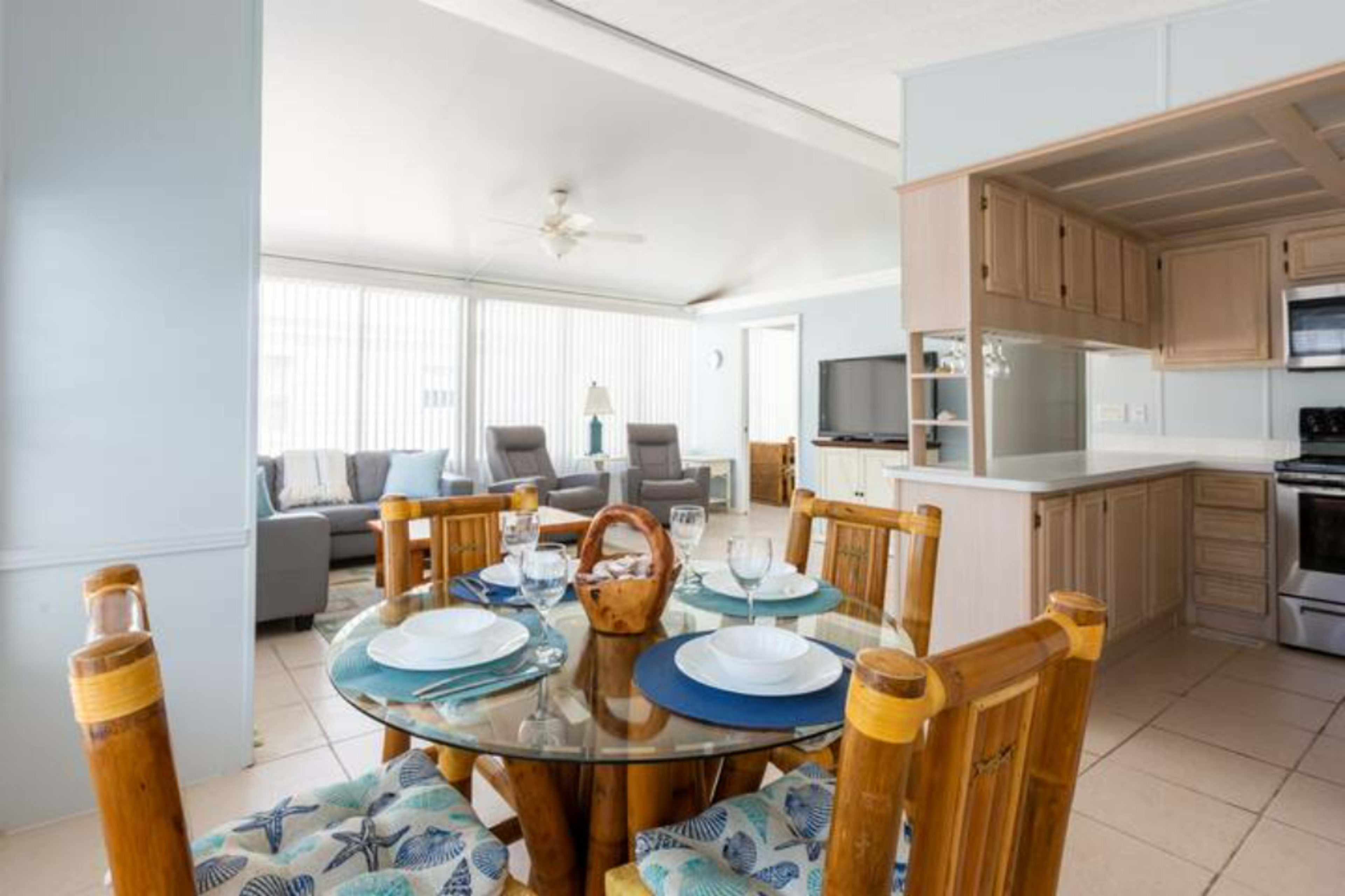 A bright open-concept living space featuring a dining area with a glass table and bamboo chairs, connected to a cozy sitting area with a couch and a television.